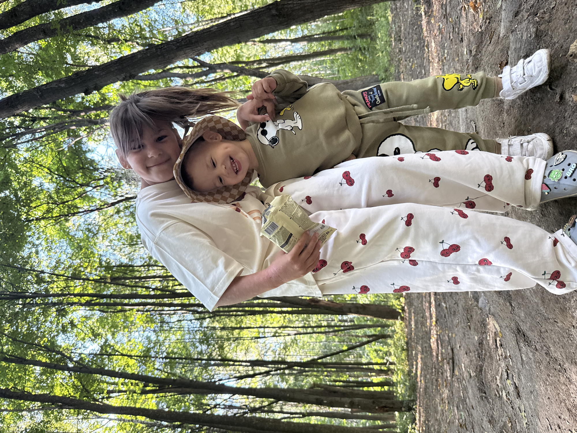 In a sun-dappled forest, two young children, a girl and a boy, are posed for a photo. The girl, on the left, has brown hair pulled back and is wearing a cream-colored t-shirt and white pants adorned with a pattern of red cherries. She holds a small, rectangular package in her right hand and rests her left arm around the boy. The boy, on the right, wears a tan, checkered bucket hat and a light green long-sleeved shirt featuring a graphic of Snoopy. He also wears matching light green pants with a Snoopy character graphic on the right leg. Both children are smiling. They are standing on a dirt path with fallen leaves, surrounded by tall trees with green foliage. The location is identified as the city of Dumino, Russia.