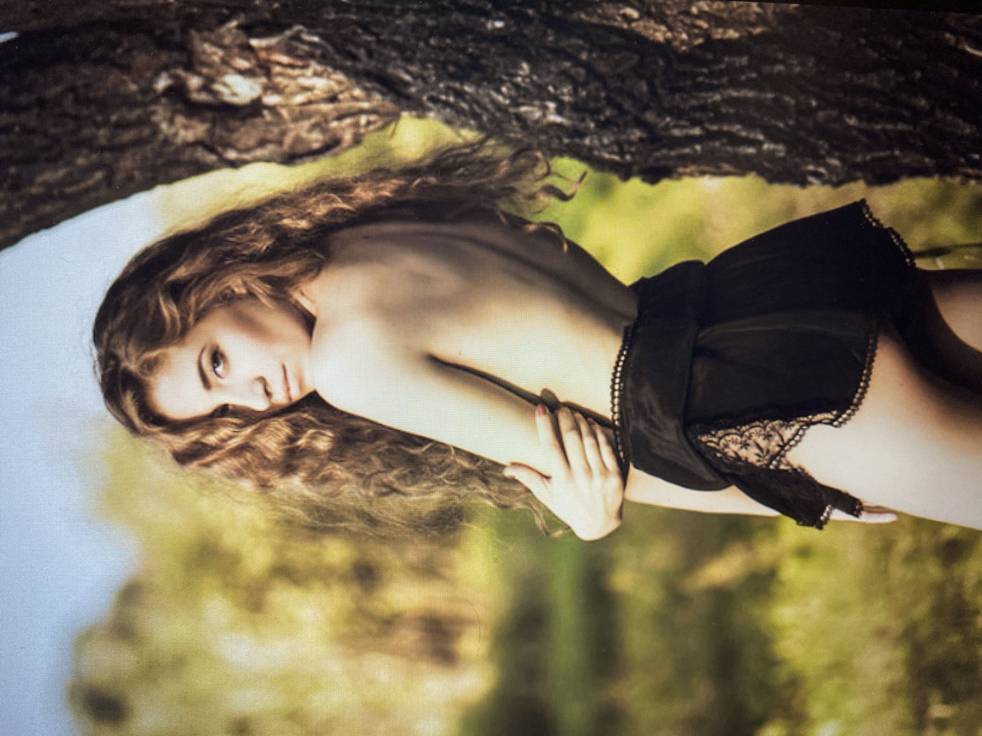 A young woman with long, curly, light brown hair stands facing away from the camera, looking over her shoulder. She is bare-chested, with her arms crossed in front of her. She is wearing black lace-trimmed shorts. She is standing next to a tree with dark, textured bark. The background is a blur of green and yellow foliage, suggesting an outdoor, possibly wooded, setting. The sky above is a pale blue.