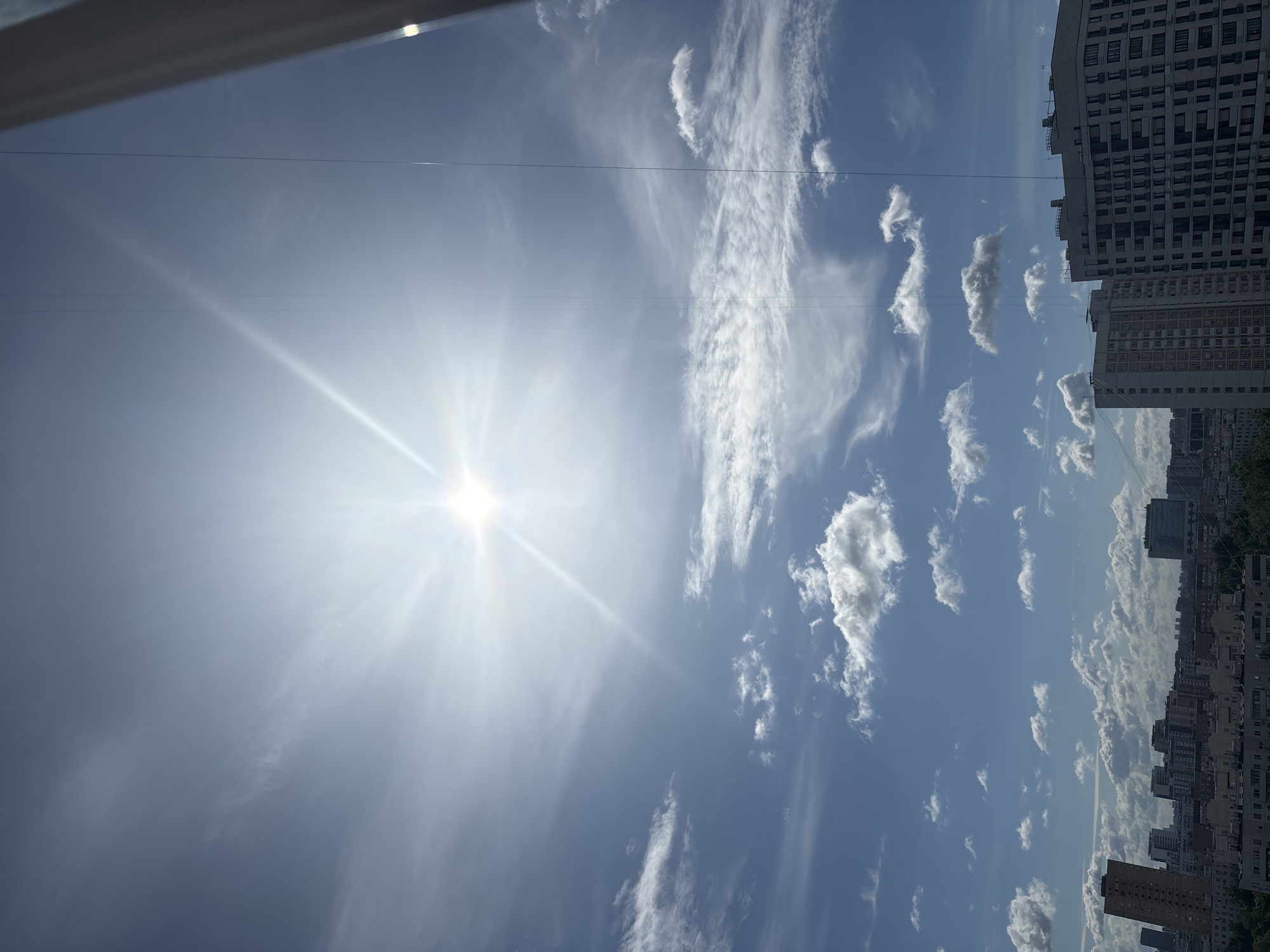 The image captures a bright, sunny sky over a city, likely Khimki, Russia. The sun is prominently featured, emitting strong rays that create a lens flare effect. Wispy white clouds are scattered across the clear blue sky. In the lower portion of the image, a cityscape is visible, with several multi-story buildings and some greenery. The perspective suggests the photo was taken from a high vantage point, possibly a balcony or window. No people or specific actions are discernible. The overall environment is urban and appears to be during daytime.