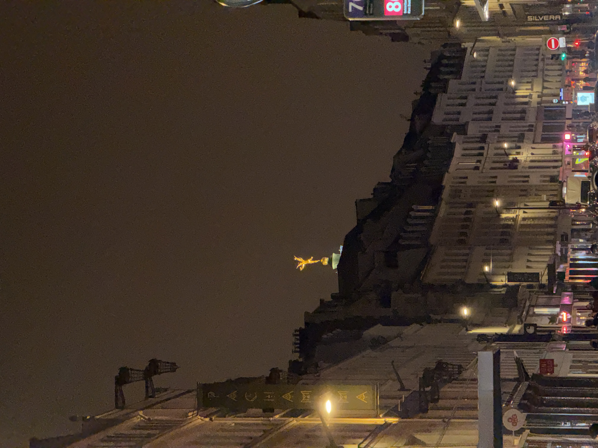 The image depicts an upside-down view of the city of Paris at night. Prominently, the gold flame atop the Pont d'Alma can be seen. Architectural details of surrounding buildings are visible, including rooftops and windows illuminated by street lights. Street signs, including one with the word "SILVERA" on it, and various neon signs indicate a busy urban street scene. There are no visible people or actions, just the static urban environment.