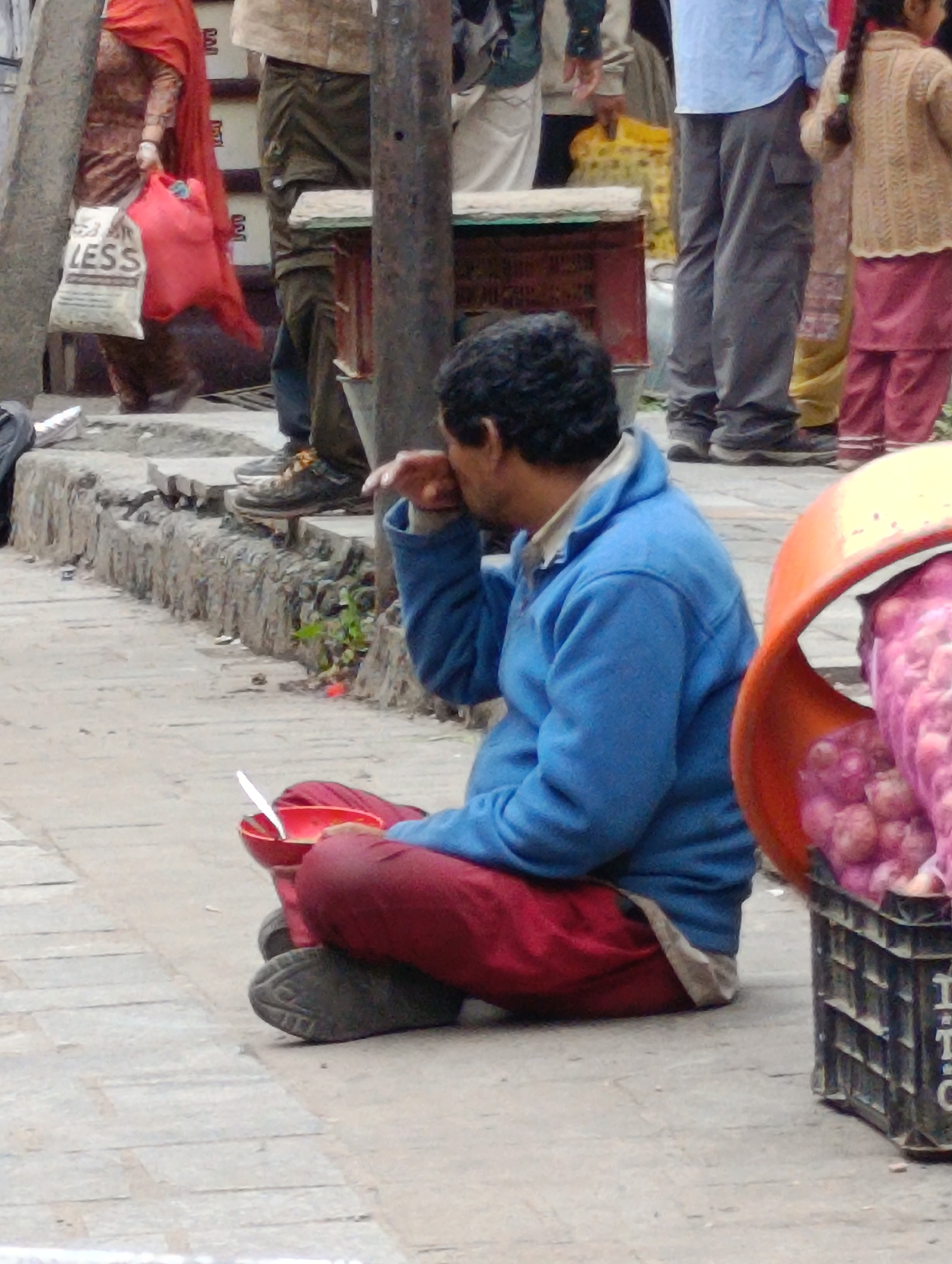 In Manali, India, a man is seated on the ground, holding a red bowl and a spoon, and appears to be eating. He is wearing a blue jacket and red pants. In front of him is a basket filled with pink onions. Behind the man, several people are visible, including a woman carrying shopping bags and a young girl. The scene takes place on a paved walkway next to a street.