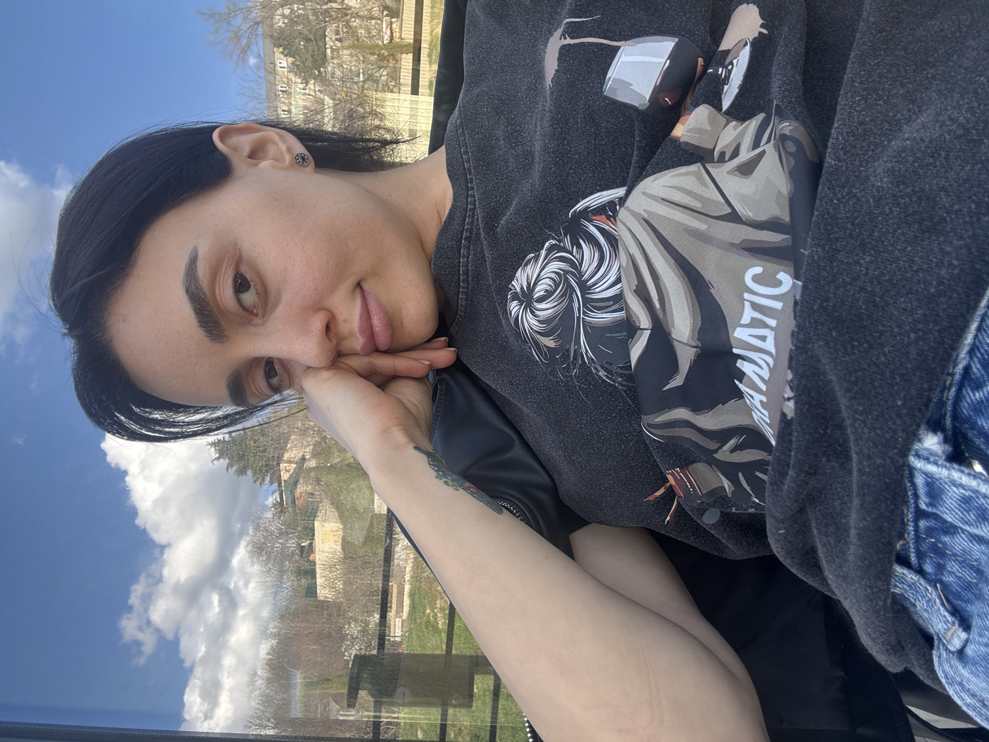 A young woman with dark hair and fair skin is positioned in a slightly tilted, close-up shot. She is resting her face on her left hand, with a relaxed expression. She is wearing a dark-colored sweatshirt with a graphic print on it and a dark jacket underneath, with a hint of a denim fabric visible. On her left forearm, there is a tattoo.
The background reveals an outdoor cityscape under a blue sky with scattered clouds. There are buildings and trees, indicating an urban environment. Based on the architectural style and vegetation, it is plausible that this image was taken in the city of Tbilisi, Georgia. The image is taken from inside a vehicle, through the window.