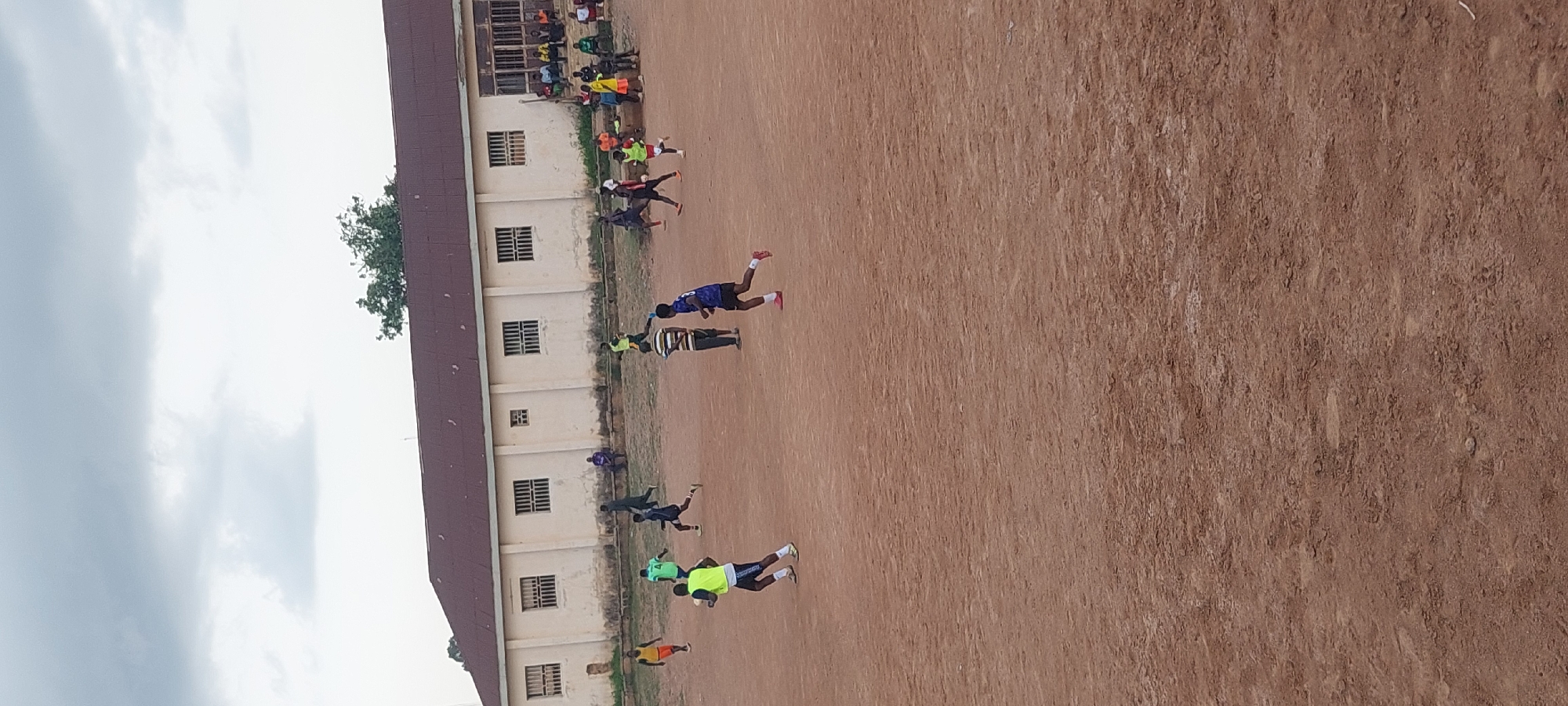 In Bukuru, Nigeria, a group of young men are engaged in a soccer match on a dirt field in front of a long, low building. The building is painted a pale yellow and has a dark red, sloped roof. Its facade features evenly spaced, barred windows and several doorways. Spectators are seated on benches along the building's exterior, some watching the game while others are engaged in conversation. The sky above is overcast with gray clouds. The field is uneven and dusty, with some patches of dry grass. The players are dressed in athletic wear, with some wearing brightly colored vests over their shirts, suggesting they are divided into teams. The overall scene depicts a casual sporting event taking place in an outdoor urban environment.