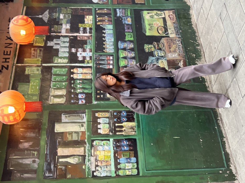 A young East Asian female stands on a grey paved sidewalk in Yuzhong District, China, positioned centrally in front of a large, colorful mural. The mural depicts the interior of a traditional Chinese store, featuring green shelving stocked with various painted items, including bottles, packaged goods, and bowls of food, with Chinese characters and "ZHENYUAN" visible on the upper section. Two illuminated orange paper lanterns with red tassels hang above the mural. The female wears an oversized dark grey bomber jacket with a lighter grey collar, a dark blue sweatshirt, and matching dark grey wide-leg trousers. She has long, dark brown hair and light-colored athletic shoes. Her head is turned slightly to the right, and her expression is neutral.
