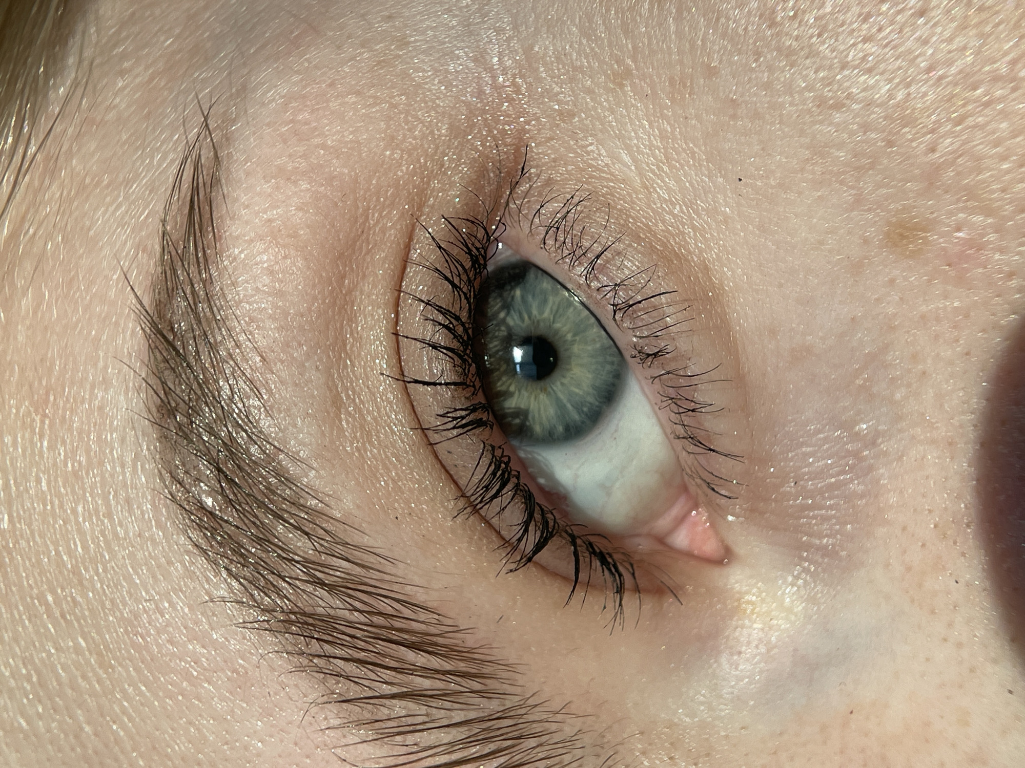 A close-up image of a human eye and eyebrow. The eye has a greenish-gray iris and visible eyelashes coated with mascara. The eyebrow is brown and appears to be well-groomed. The skin around the eye shows pores and slight imperfections, with a few visible freckles. The sclera (white part of the eye) is slightly red. The background is out of focus. There is no information in the image suggesting its location is Izhevsk, Russia.