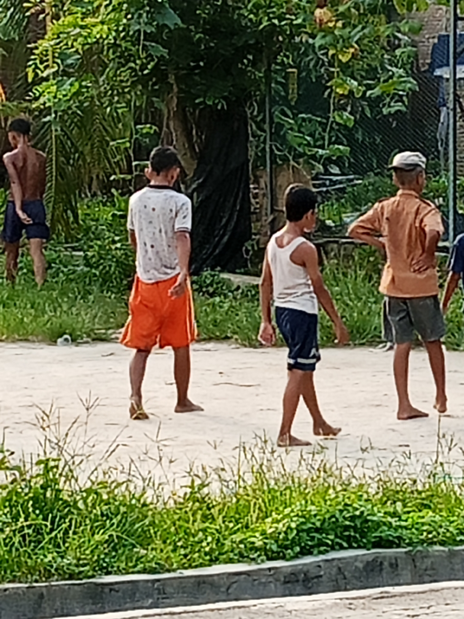 In Bandar Labuhan, Indonesia, four young males are seen in an outdoor setting. On the far left, a bare-chested individual with dark hair is wearing blue shorts. Next to him, a boy in a white t-shirt with small dark dots and orange shorts is facing away from the camera. To his right, another boy in a white tank top and blue shorts with white stripes is walking. The furthest person to the right is a boy wearing a light brown collared shirt and grey shorts, with a white cap on his head, facing away. The ground appears to be dirt or sand, with patches of grass and foliage in the background. A chain-link fence is visible behind the individuals.