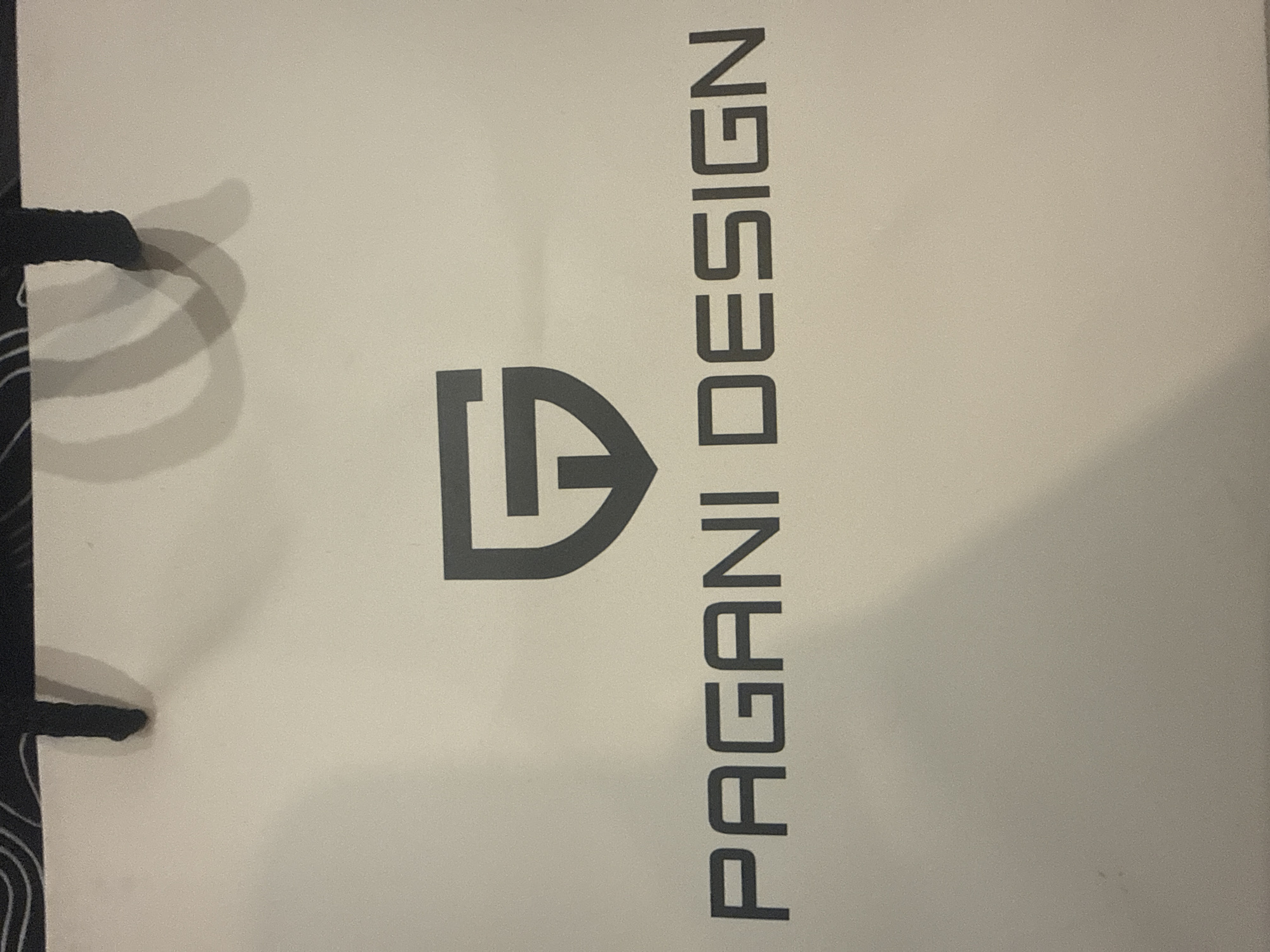 A white paper shopping bag with the logo and name "PAGANI DESIGN" printed in dark gray is displayed. The logo is a stylized "G" within a shield shape. The bag appears to be a product packaging item. The image is cropped to show only the bag and its branding, with the top portion of the bag showing the black handles. There is no visible context or indication of its location in Burgas, Bulgaria.