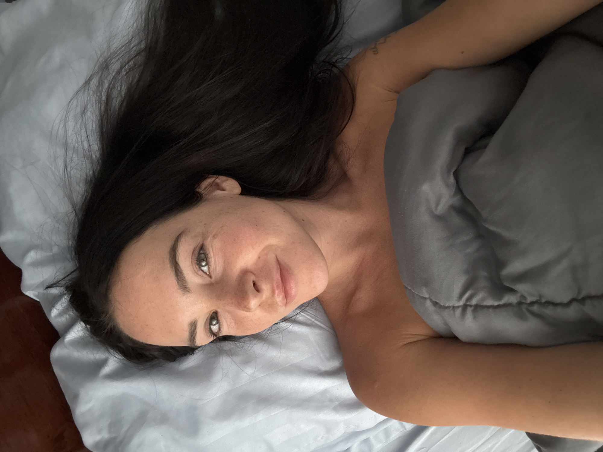 A woman with dark hair and fair skin with freckles lies in bed, looking at the camera. She is partially covered by a gray comforter. Her hair is spread out on light blue sheets. A dark wooden headboard is visible at the top of the image. The scene is an indoor bedroom.