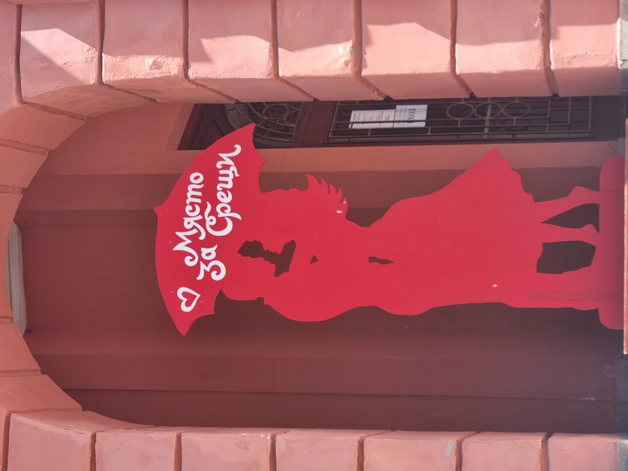 A red sign is mounted on a wall, partially framed by a peach-colored brick arch. The sign is in the shape of an umbrella with the words "Място За Срещи" written vertically in white. A white heart shape is situated beneath the first word. The silhouette depicts a couple standing under the umbrella. Above the sign, through the arch, is a black metal grate with oval cutouts. Above the metal grate, there is a white sign with black lettering, partly obscured. The image was taken in the city of Pleven, Bulgaria.