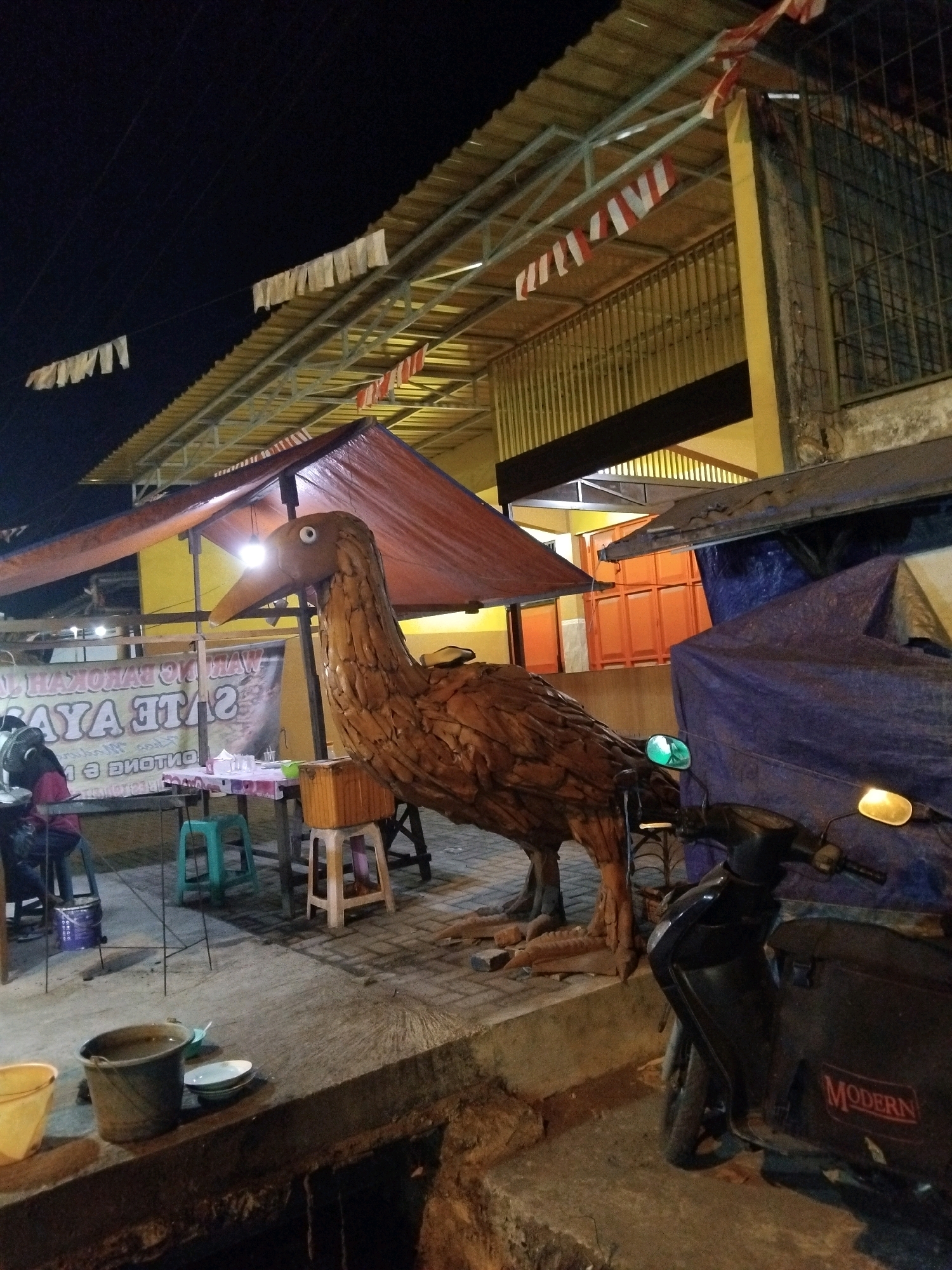 In Wedoro, Indonesia, at night, a rustic wooden sculpture of a large bird stands prominently on a paved area. The bird is brown, with visible segments forming its body and neck, and a distinct eye and beak. To the left of the bird, a red tarp is stretched over a frame, creating a makeshift shelter under which tables and chairs are set up. A banner with text is visible behind the tables. A person wearing a pink shirt and black pants is seated at one of the tables, with a fan visible to their left. A motorcycle, partially covered by a blue tarp, is parked to the right of the sculpture. The background shows a building with a yellow facade and red-orange doors, beneath a sloped roof adorned with white and red banners, suggesting a festive occasion. The ground is paved with light-colored stones, and a dark trench runs along the bottom of the frame. Various containers, including buckets and bowls, are scattered on the ground near the tables and in the foreground. The overall scene depicts an outdoor gathering or market area at night.