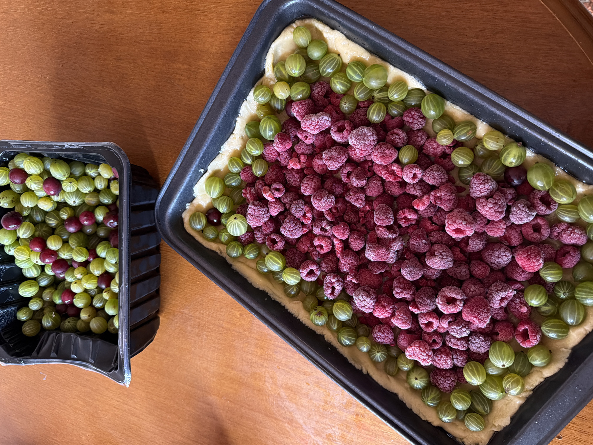 Two black plastic containers of berries are on a wooden surface. In the upper container, there are numerous green and red gooseberries. In the lower container, which is rectangular, there is a crust with a filling of raspberries in the center and a border of green gooseberries. The scene is a close-up shot, taken from a high angle. There is no indication of the location in Moscow, Russia.