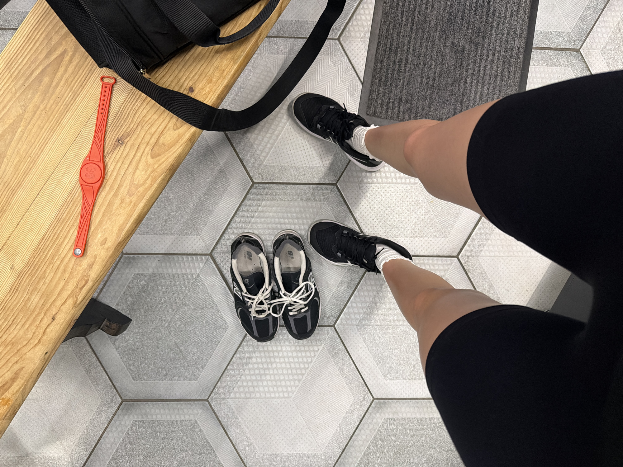 The image shows a pair of black athletic shoes and a person's legs and feet. The shoes are on a floor with hexagonal tiles. One shoe is in the process of being put on, with a white sock visible above the shoe. The other shoe is lying next to it. A black bag is on a wooden bench in the background. A red wristband is also on the bench. The scene appears to be a locker room or a gym. The lighting is neutral, suggesting it could be any time of day. There is no visible text in the image. The overall mood is functional and perhaps preparatory for an athletic activity.