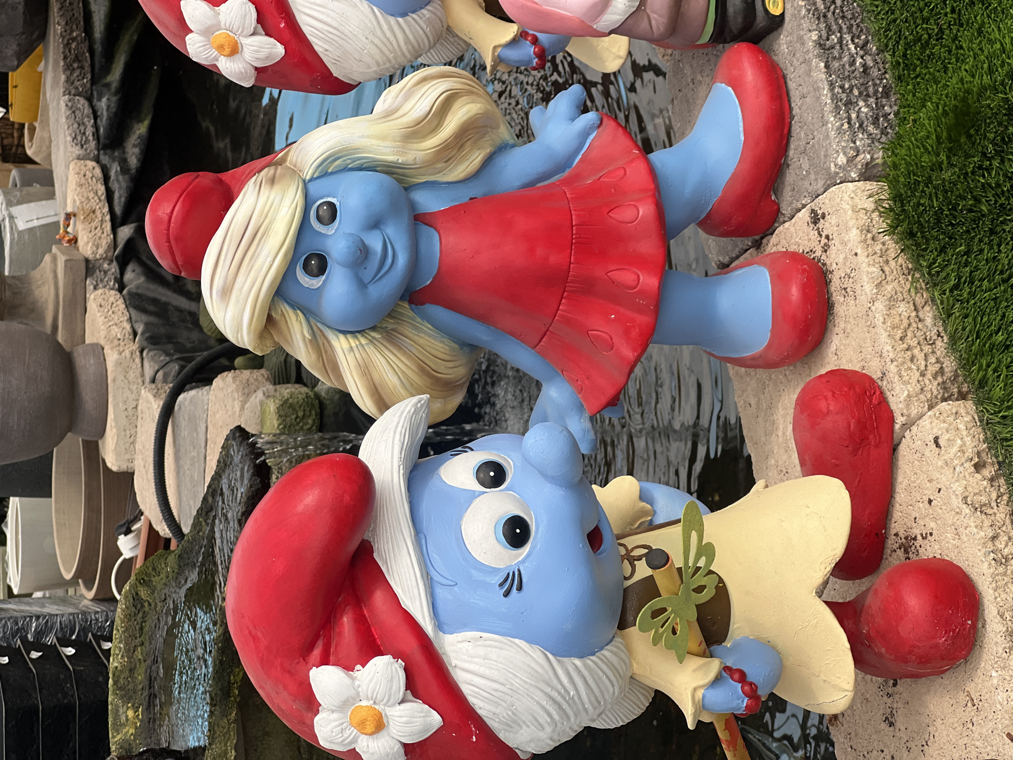 This image shows a collection of Smurf figurines, likely garden ornaments, displayed near a water feature. One figurine is Smurfette in a red dress, while another is a male Smurf with a white beard, red hat with a white flower, and a musical instrument. The figurines are positioned near a stone-like structure, with a glimpse of green grass visible in the background. The scene appears to be outdoors, possibly in a garden or a retail setting selling garden decorations. There is no information present to determine the location as Zvulun Regional Council, Israel.