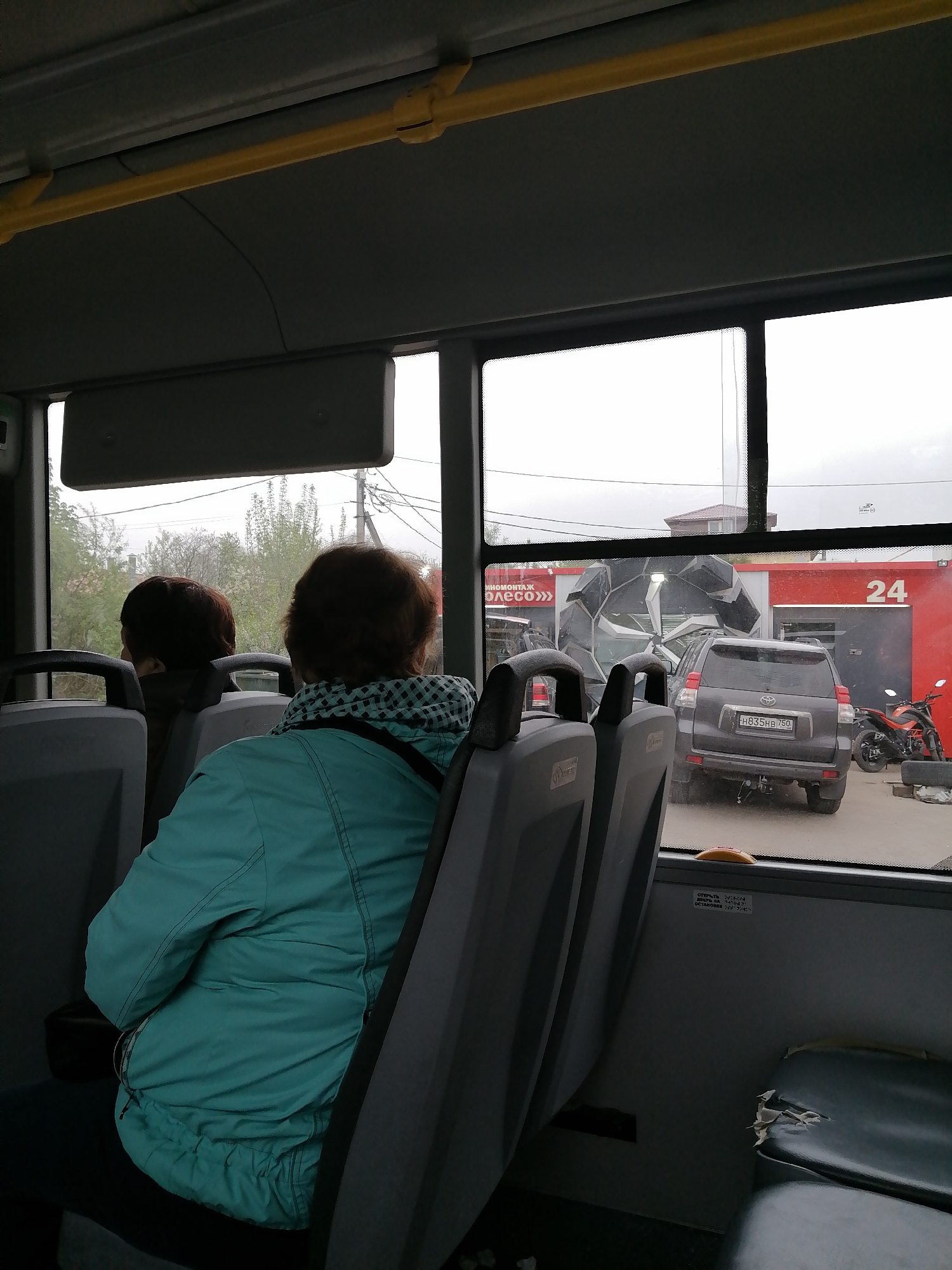 From the interior of a bus, a rear-view shot of two women seated, with a view through the window of a gray SUV parked outside a building with a red facade marked with the number 24. Beside the SUV is an orange motorcycle. An abstract metallic structure is visible behind the SUV. The location is identified as an area in the city of Unknown, Unknown. The bus interior shows gray plastic seating, yellow overhead handrails, and other standard bus fixtures.