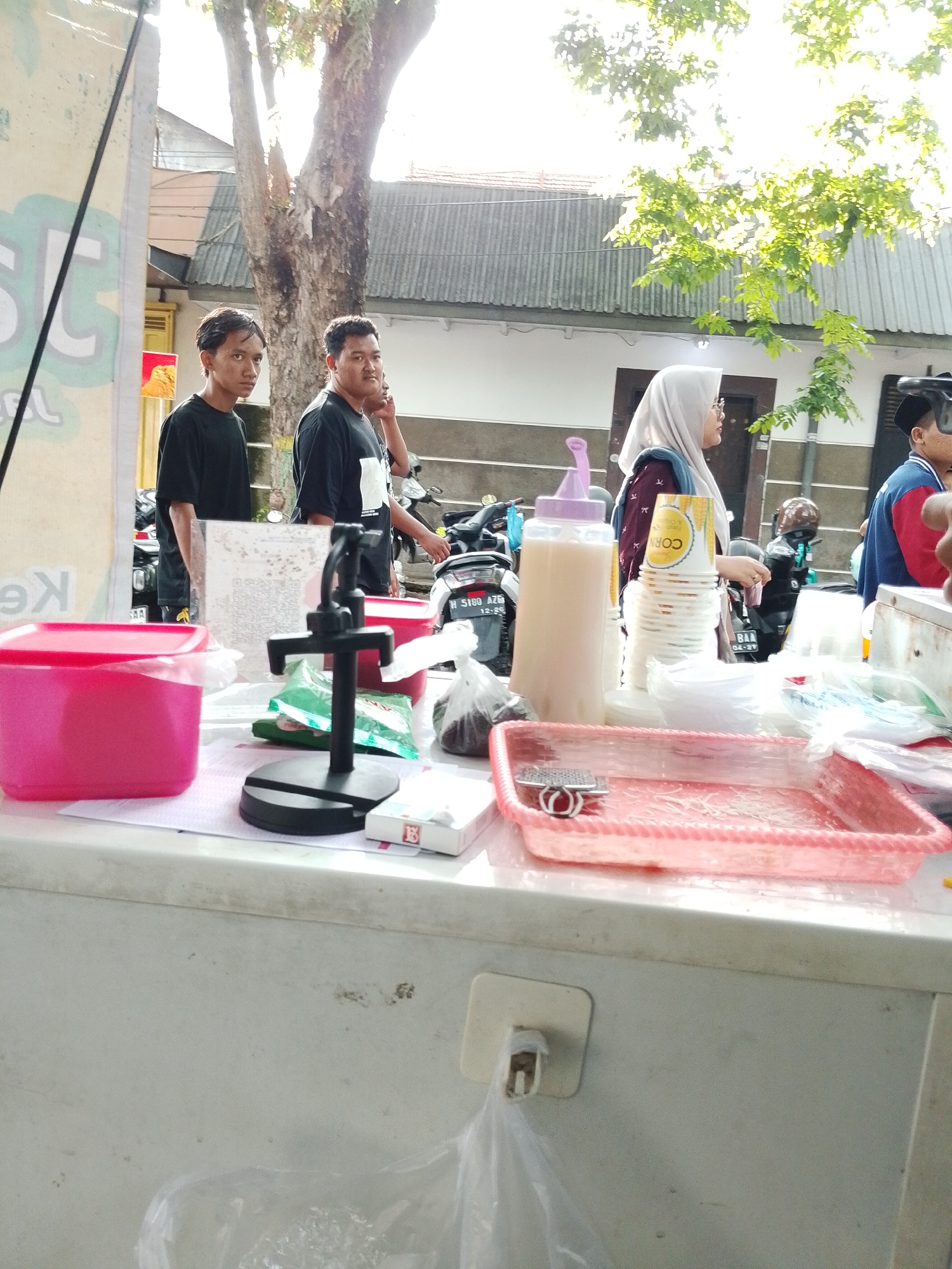 Scene: An outdoor food stall in Kecamatan Pati, Indonesia.
People: Two young men stand near the stall. A woman wearing a headscarf stands at the stall, presumably purchasing food. A person in a blue and red top is partly visible behind her.
Objects: A variety of containers are on the stall counter, including pink plastic tubs and a bottle with a purple lid. Also present are cups, plastic bags, a cash register stand, and a pack of cigarettes.
Actions: People are standing at the stall; likely there is a food transaction taking place.
Interactions: Possible customer/vendor interaction.
Environment: The stall is set up on a street, with motorbikes parked in the background. A tree is visible to the left of the stall. The weather appears bright and sunny.