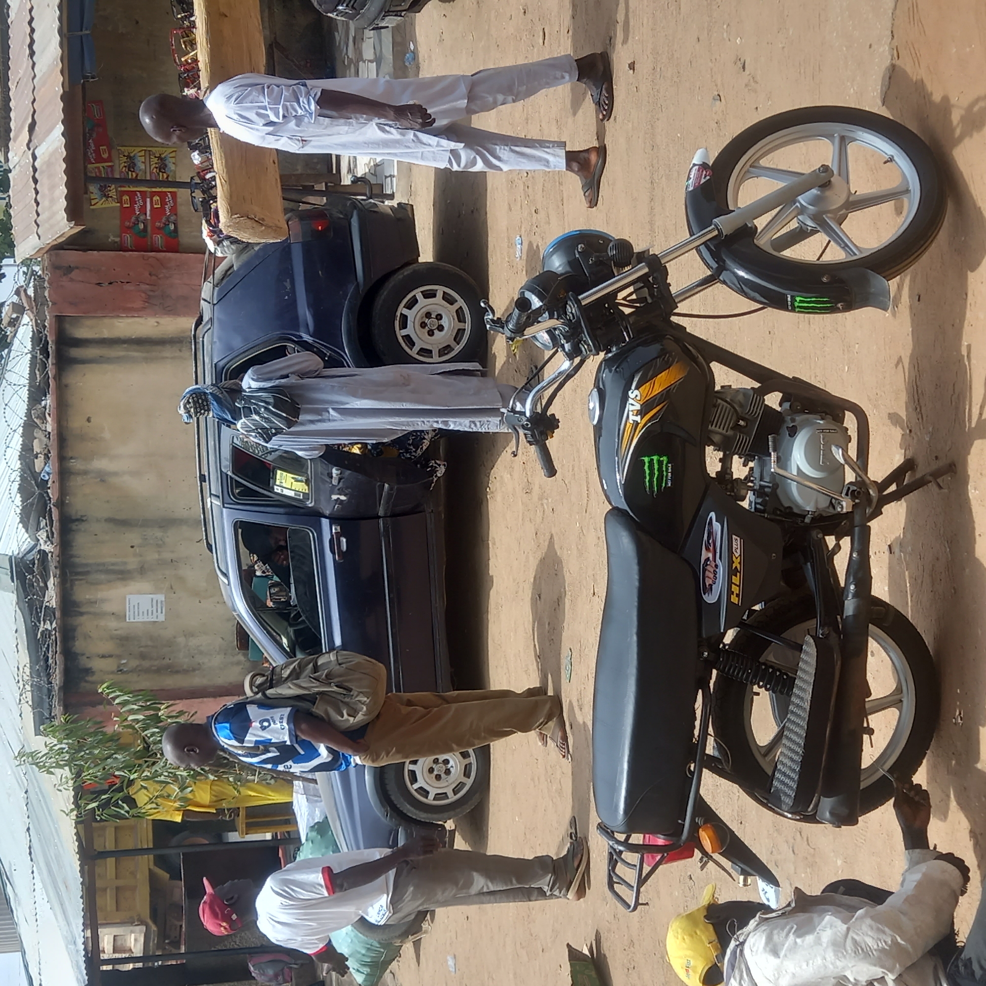 Dadi Riba, Nigeria: A black TVS HLX Plus motorcycle stands on a dirt road, partially obscuring a blue car. Several individuals are present: one man in a white robe stands near the car's open door, another man in white stands to the left of the car, one man in blue stands to the left of the car and in front of one man in white wearing a red cap and one man in light colors and a yellow cap. The surroundings include buildings with corrugated metal roofs, a shop front displaying beverage advertisements, and a wall topped with barbed wire.