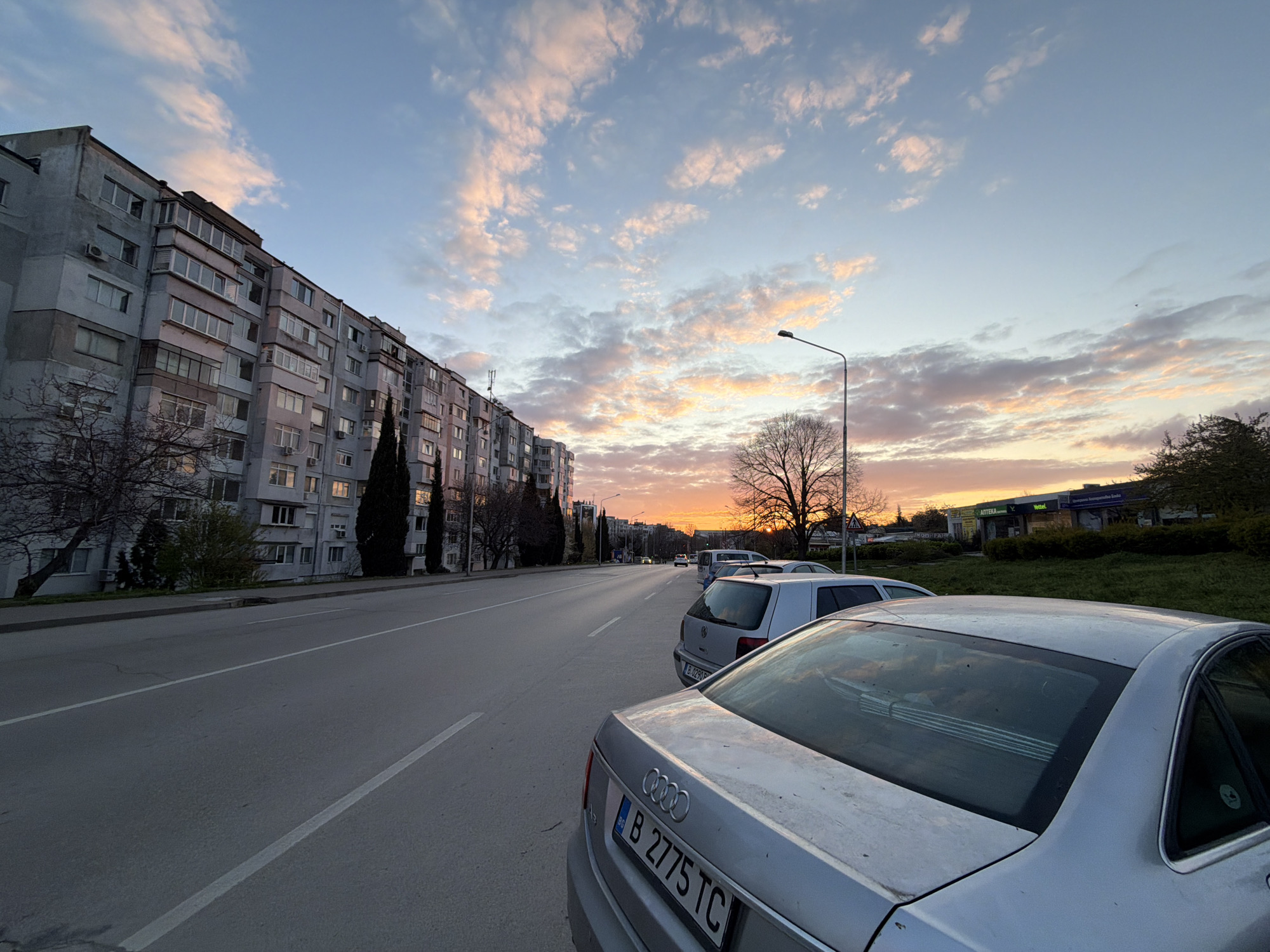 The image shows a street scene in Varna, Bulgaria at sunset. A long, grey apartment building lines the left side of the street. The street is wide and asphalted, with a white dividing line. Several cars are parked along the right side of the street, including a silver Audi in the foreground with the license plate "B 2775 TC". In the background, the setting sun casts a warm glow on the horizon and colors the clouds pink and orange. A lamppost stands on the right side of the street. On the far right of the street, there is a green grassy median, with buildings that have signs reading "Аптека" and "Vettel" visible in the background. There are no pedestrians visible in the image.
