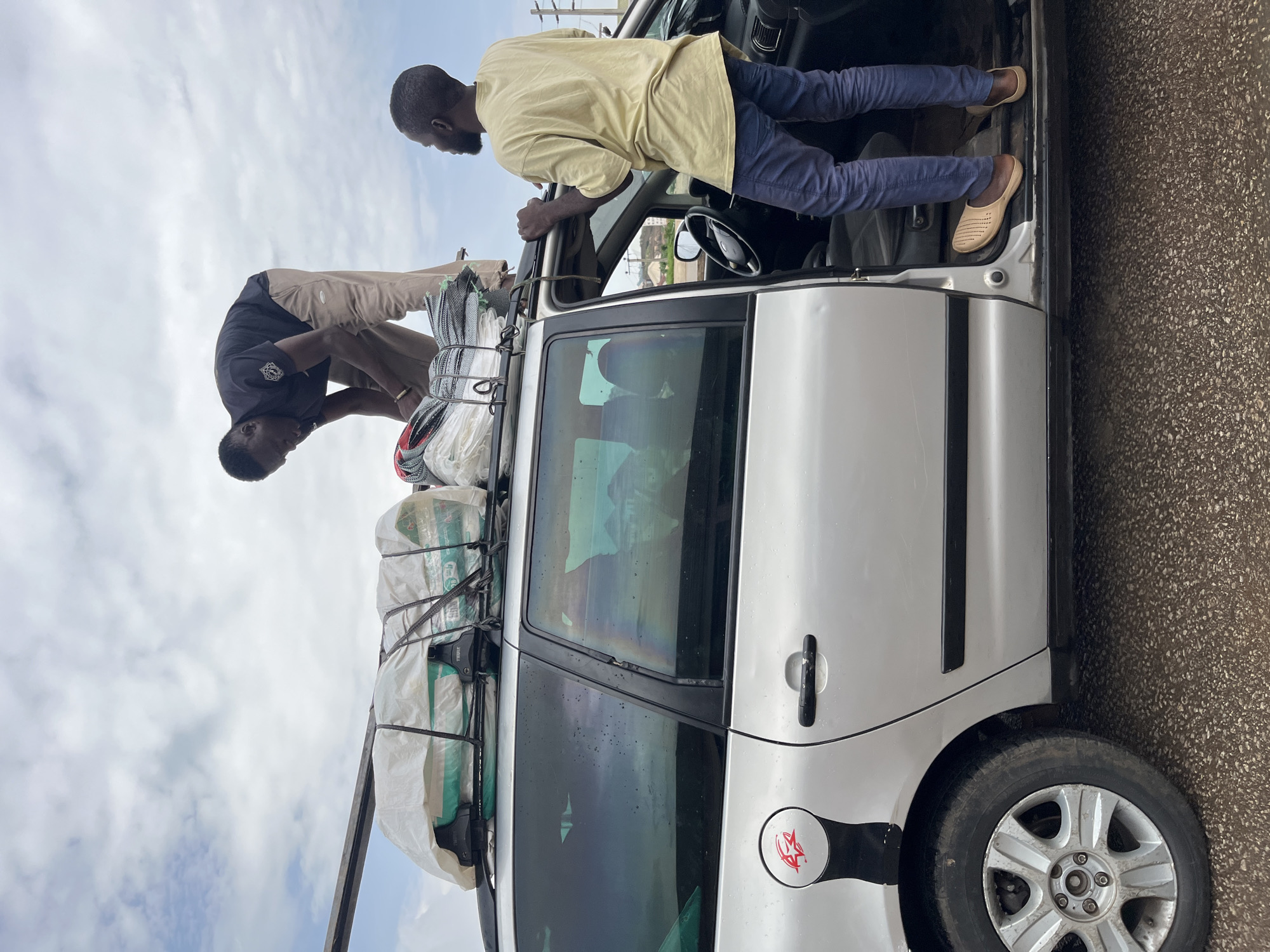 Two men are seen loading bags onto the roof of a silver station wagon, which is parked on a paved road. One man, wearing a black t-shirt and tan pants, is standing on the roof of the car, adjusting the bags. The other man, wearing a light yellow t-shirt and blue pants, is standing with the car door open, his feet on the running board, and his hands on the door frame. The bags on the roof are wrapped in white plastic and secured with black straps. The car has a roof rack. The background shows a cloudy sky and some distant buildings and utility poles, suggesting an urban or semi-urban environment. The location is identified as Gura Top, Nigeria.