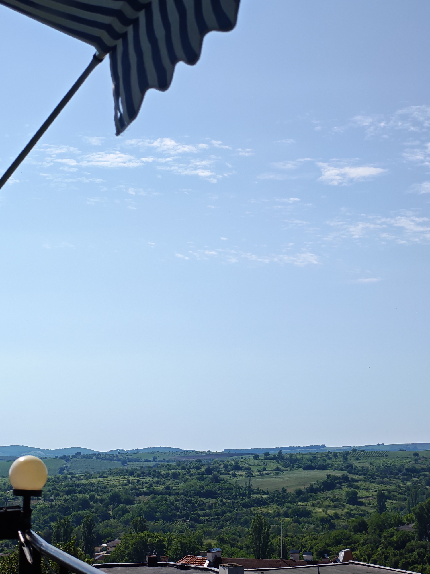 The image presents an elevated panoramic view of a rural landscape under a clear, light blue sky with scattered white cumulus clouds. In the upper left quadrant, a segment of a dark blue and white striped fabric awning or umbrella, supported by a dark pole, is visible. The immediate foreground, at the bottom of the frame, features the dark rooftops of residential or commercial buildings. A spherical, yellowish-white outdoor lamp, attached to a dark metallic railing, is prominently positioned in the lower left. Beyond the rooftops and railing, the midground is dominated by a vast expanse of verdant rolling hills and valleys, densely covered with various species of trees and shrubs in multiple shades of green. Interspersed within this greenery are visible patches of lighter green fields or agricultural land. In the distant background, gentle hills with a slightly hazy, blue-green tone meet the horizon line. No human figures or large animals are discernible; however, a few tiny dark specks, possibly birds or insects, are visible against the sky. The scene is situated in the city of Ivaylovgrad, Bulgaria.