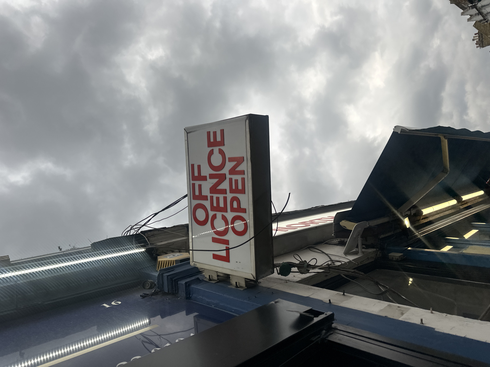 A low-angle shot reveals an "OFF LICENCE OPEN" sign, vertically oriented and predominantly white with red lettering. The sign is mounted on a building's exterior, surrounded by an array of electrical wires and assorted fixtures. The facade of the building features a blue-painted surface with the number "16" visible. Above the sign, sections of the building's structure and lighting fixtures are apparent. The background is dominated by an overcast sky. The location is the city of London, United Kingdom.