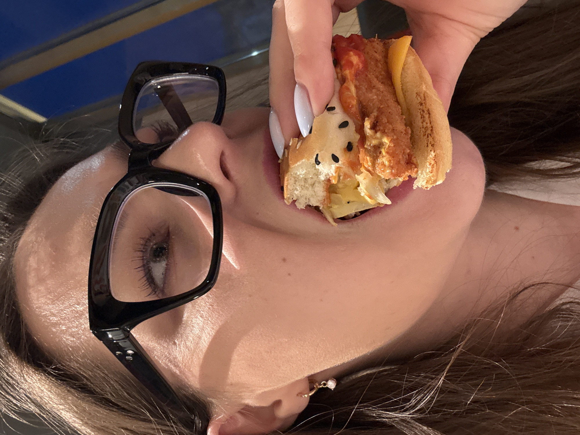 A close-up, angled shot captures a person, likely female, eating a small hot dog or mini sandwich. The person is positioned with their head tilted upwards, possibly lying down. They wear black, thick-rimmed glasses, reflecting ambient light, and have visible light brown hair. A delicate gold earring with a white charm is seen on their right earlobe.
A hand with long, white, almond-shaped fingernails holds the food item towards the person's open mouth. The food consists of a toasted bun filled with what appears to be a fried sausage or piece of chicken, melted yellow cheese, a red sauce (possibly ketchup), and shredded light-colored vegetables. Black seeds are sprinkled on the bun, which has a visible bite taken from it.
The background is out of focus, showing a dark blue area, possibly a night sky or a dark room, with some faint horizontal lines that could be a railing or window frame. The lighting indicates an indoor setting, likely in the evening or at night. There is no visible text in the image. The provided context for the image's location is Izhevsk, Russia.