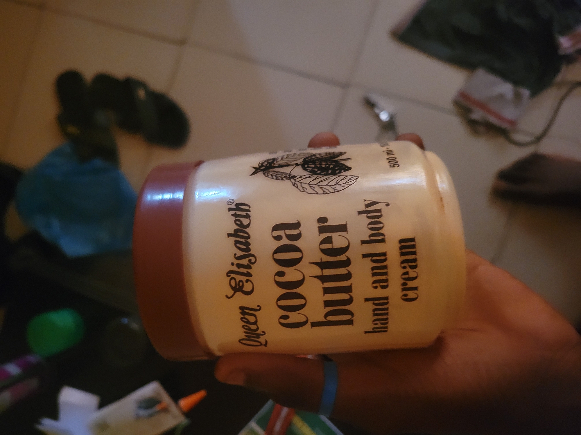 A person's hand, wearing a blue bracelet, holds a jar of Queen Elisabeth Cocoa Butter hand and body cream. The jar is off-white with a brown lid and features black text and an illustration of cocoa pods. The background is blurry and appears to be an indoor setting with various objects.