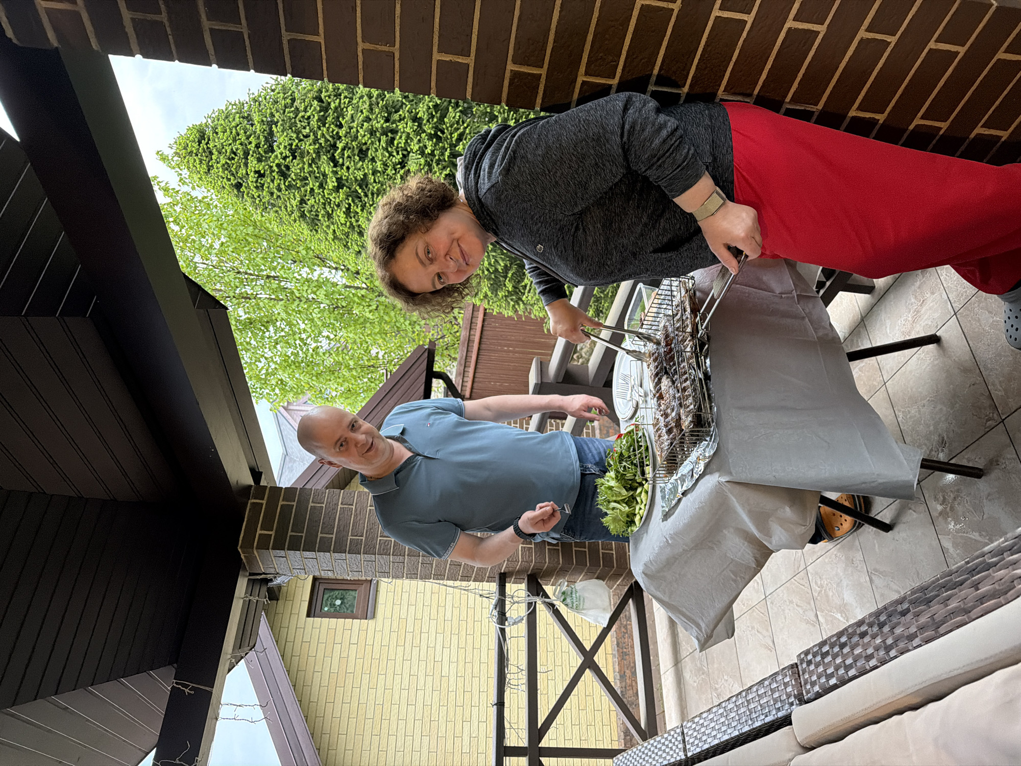 A woman in a gray hoodie and red pants is grilling food on a grill covered with aluminum foil. She is using tongs. A man in a blue polo shirt and jeans is standing next to her. There is a table next to the grill with a plate of salad. The scene is outdoors, on a patio with a brick column and a view of trees. The location is Новоглаголево, Russia.