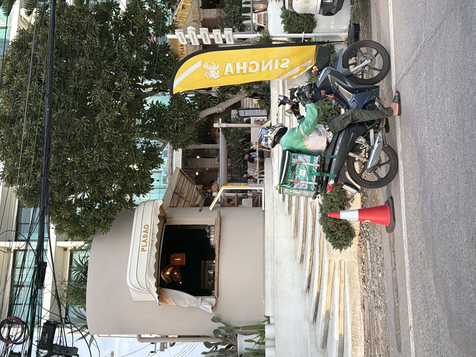 Here is a description of the image:
A sideways outdoor daytime shot depicts a street scene in Pattaya City, Thailand. On the left is a restaurant named PLADAO CAFE, with a white awning, a tan exterior, and windows through which light fixtures can be seen. A series of overhead wires are visible to the left of the restaurant, along with an electrical box. A large tree with dense foliage obscures part of the restaurant.
Next to the restaurant is a sign displaying the various goods that can be bought. Next to this is a yellow flag for “SINGHA”. A delivery person is seated on a black motorcycle parked on the sidewalk next to the restaurant, their feet resting on the street. A green delivery bag with the word “M” in white can be seen on the back of the motorcycle. The person is wearing a green long-sleeved shirt and a helmet. A red and white traffic cone is positioned on the sidewalk adjacent to the motorcycle. The rest of the frame is filled with the grey concrete street.