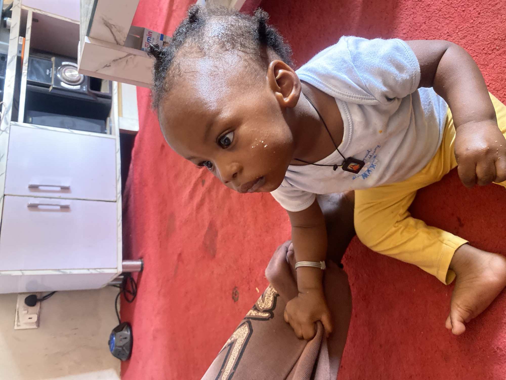 A child, approximately one year old, lies on a red carpet in a room in Rido, Nigeria. The child wears a white shirt, yellow pants, and a dark necklace. They are reaching for a cloth held by someone out of frame. A white cabinet with silver handles sits nearby. A power outlet and a blue power adapter are visible on the wall.