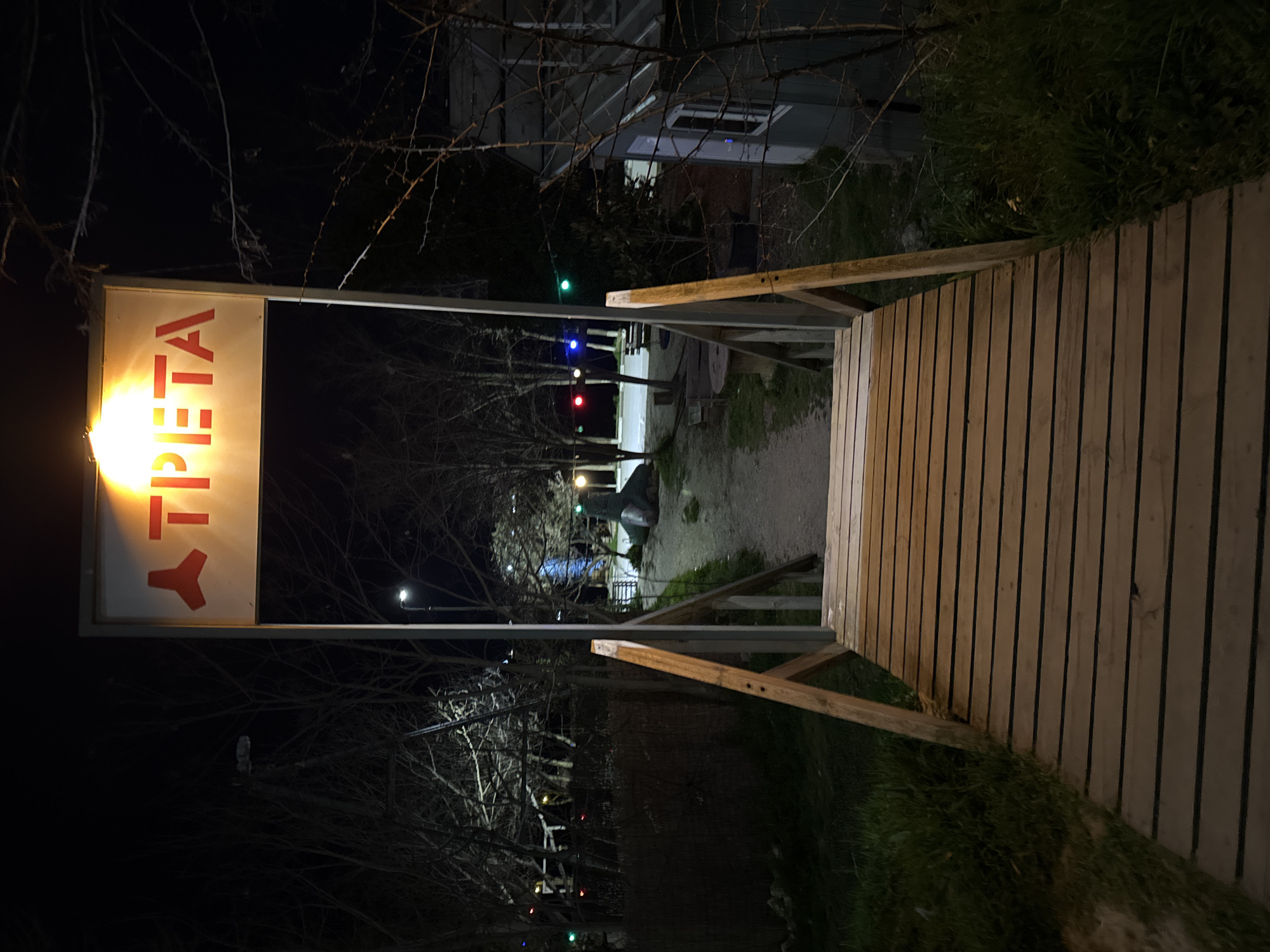 The image, taken at night, depicts a wooden bridge leading to a path. To the left of the bridge is a sign illuminated from behind, displaying the word "TPETA" vertically in red letters. The path is dimly lit, with scattered lights in the background. Bare trees line the path. A building is visible behind the bridge, with a partially visible window. The location is Varna, Bulgaria.