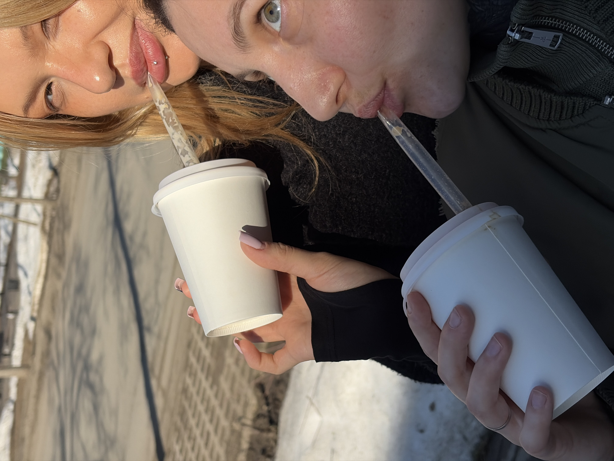 Two women are drinking from white paper cups with straws. The woman on the left has blonde hair and a lip piercing. The woman on the right has green eyes. The scene appears to be outdoors on a sunny day. The location is inferred to be Chaikovsky, Russia, based on the image request. The background includes a sidewalk and possibly a street or building, all bathed in sunlight.