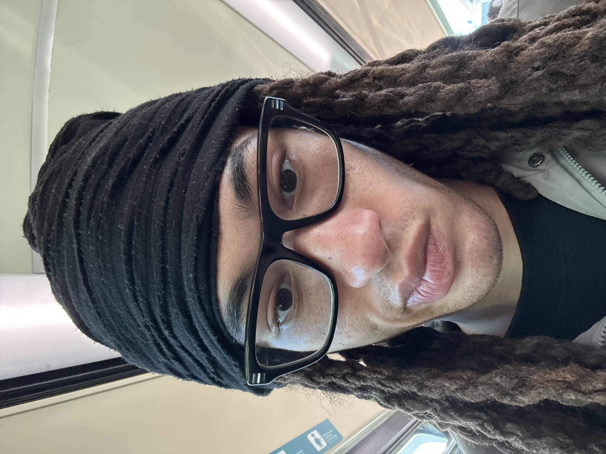 A close-up, portrait-oriented selfie features an individual with medium-dark skin, looking directly at the camera. The person is wearing a black beanie or headwrap that covers the top and left side of their head, with long, dark dreadlocks visible around the face, particularly on the right. Black-framed glasses are worn, covering the eyes. The image focuses on the face, showing the nose, full lips, and stubble around the mouth and chin. The expression is neutral and direct.
The person is dressed in a light-colored jacket with a visible zipper and a snap button on the right side, layered over a dark shirt. The background is a brightly lit, light-colored interior, likely a ceiling or upper wall, with linear light fixtures or window frames suggesting an indoor setting.
The scene appears to be on public transportation, such as a train or metro, which aligns with the provided location context of Orsay, France. A blue sign with white pictograms, possibly indicating transport rules (e.g., luggage icon, seating icon), is partially visible in the lower right, but any accompanying text is unreadable. The image captures a casual moment in transit.