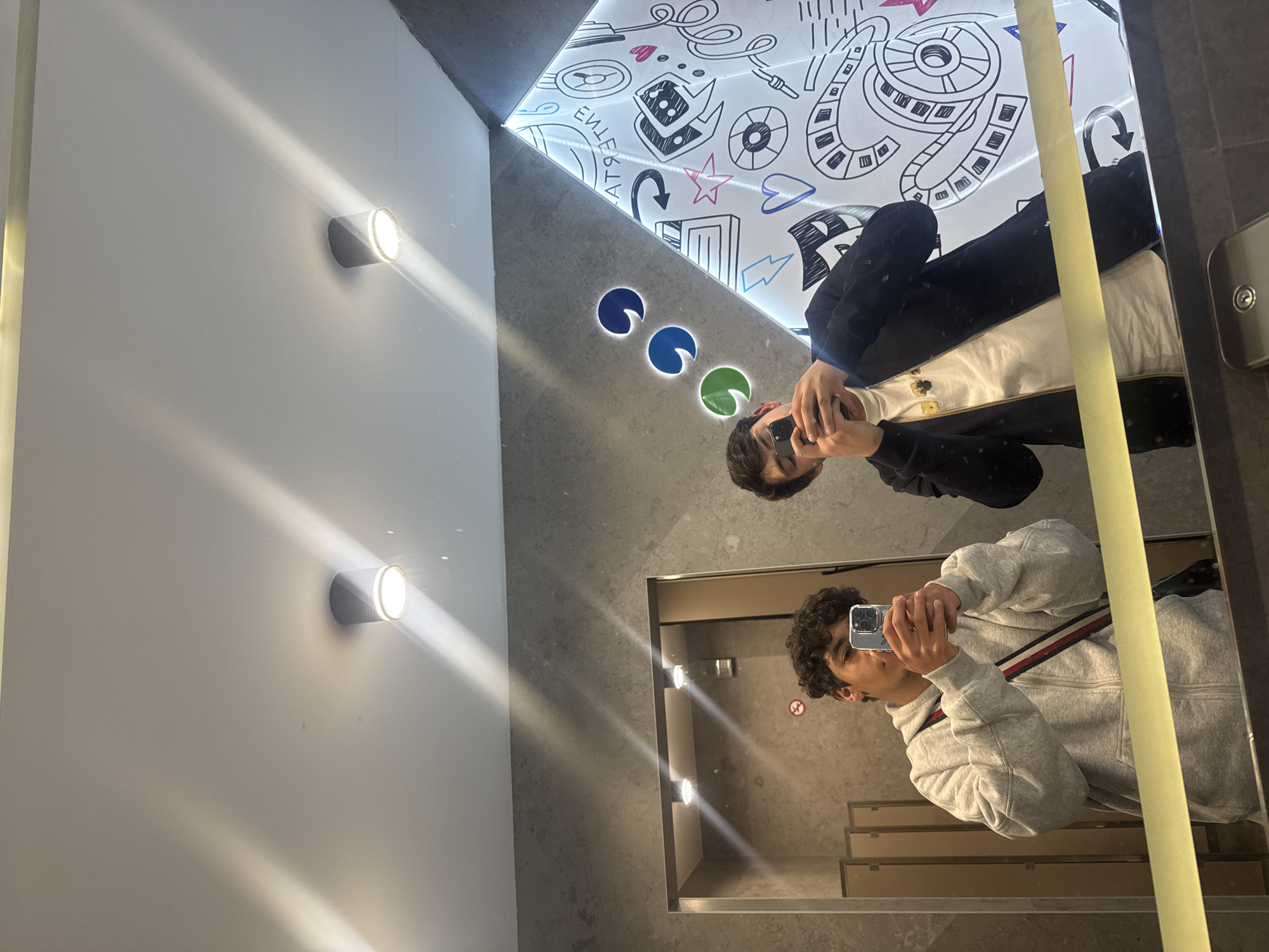 A person is taking a selfie in a restroom that has been captured in a mirror. The mirror is located in Yerevan, Armenia. The person is reflected in the mirror, facing the camera and holding a smartphone. The person's reflection shows them wearing a gray sweatshirt with a red and blue stripe. Above the person in the mirror, another reflection shows someone in a black jacket over a white shirt.
The restroom is brightly lit with cylindrical wall-mounted lights. The walls are a mix of white and grey, with a decorative ceiling featuring cartoon-like drawings and colored circles. The floor is tiled. A door frame is visible in the mirror.