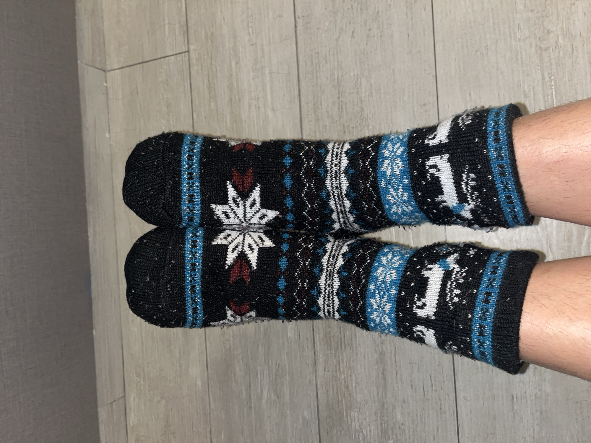 A pair of legs wearing black knit socks with a winter-themed pattern are presented on a wooden floor. The socks are black with a white snowflake design near the toes and white reindeer near the ankles. A blue patterned stripe runs vertically along the front of each sock. The legs are Caucasian. The background consists of a grey wooden floor and a grey wall. The image does not contain information to ascertain the location in the city of Buenos Aires, Argentina.