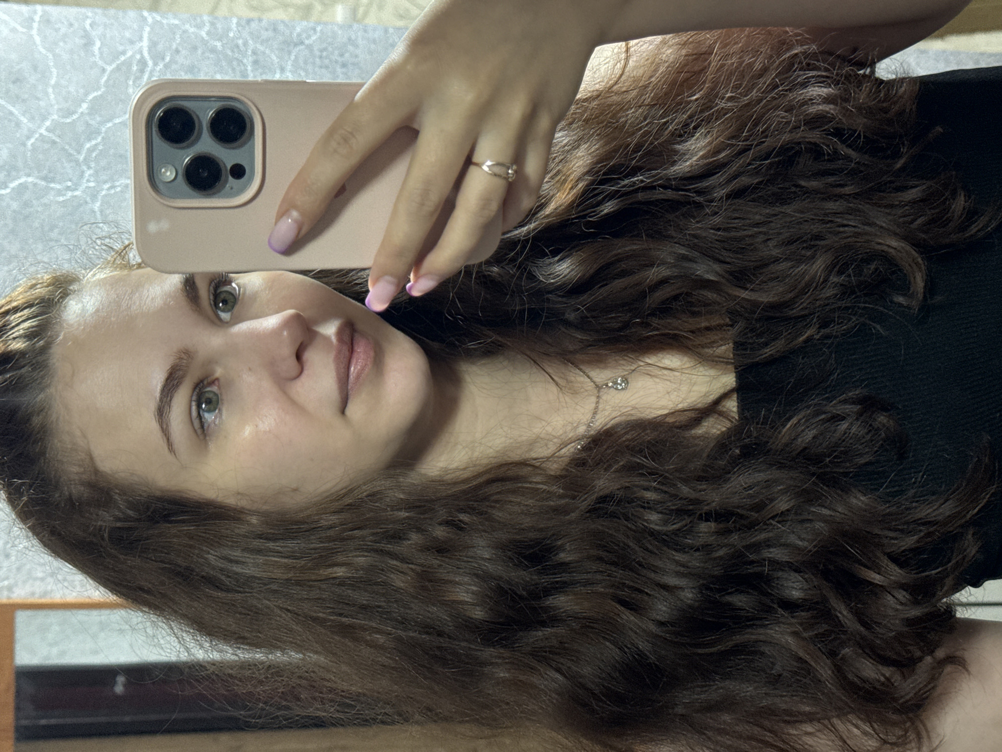 A young woman is taking a selfie in a mirror with an iPhone. She has long, wavy brown hair, fair skin, and green eyes. She is wearing a black top and a silver necklace. On her finger, she is wearing a ring. The iPhone has a pink case. Her nails are painted with a lavender french manicure. The location is unknown, and there is no evidence of it being in the city of Nizhnyaya Gar', Russia.