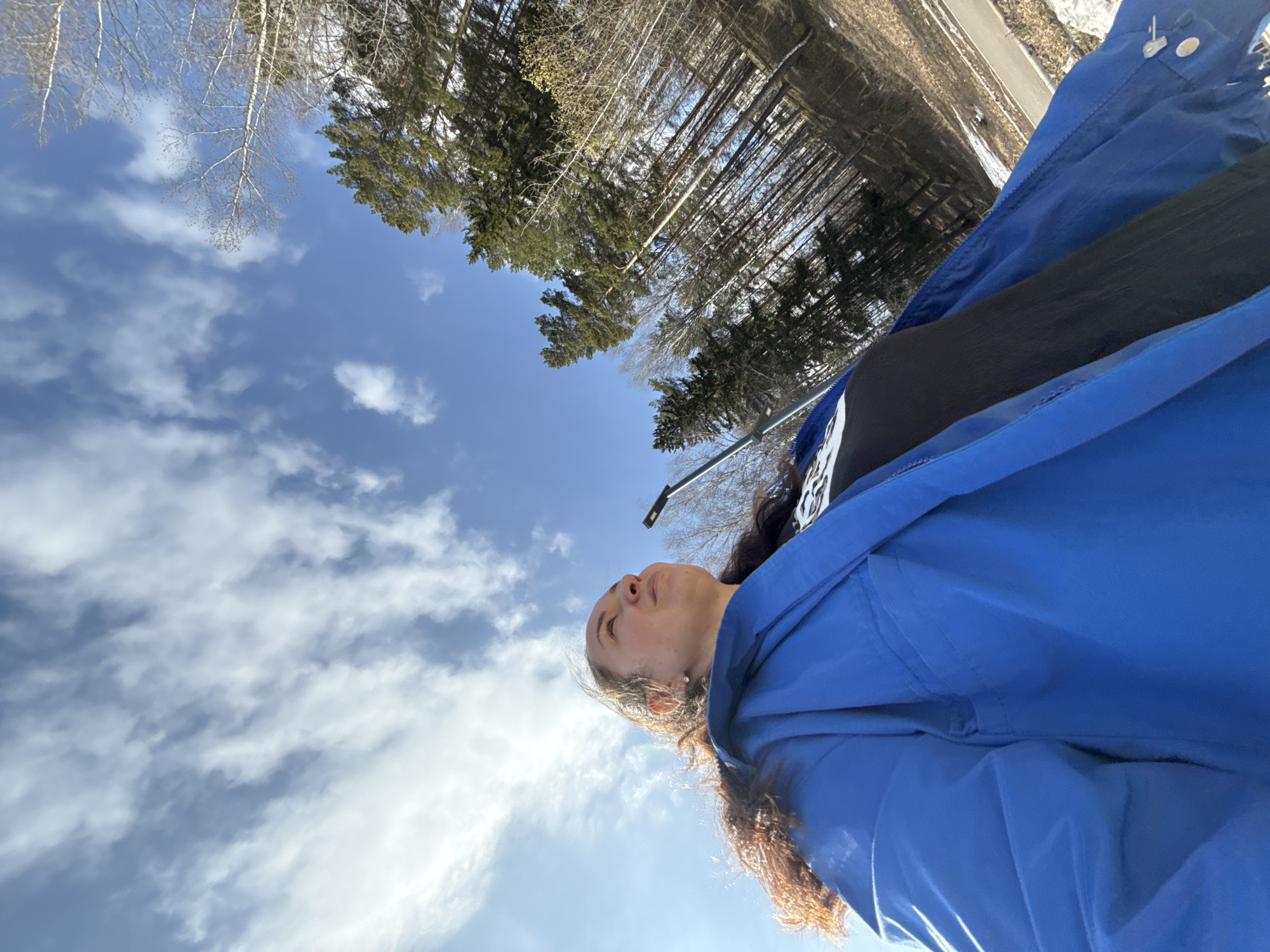 A low-angle shot depicts a woman in a blue jacket looking up at a partly cloudy sky. Some bare trees and evergreen trees are visible in the background. A road or path can be seen in the upper right of the image. It is difficult to confirm the location is Chaikovsky, Russia based on the image.