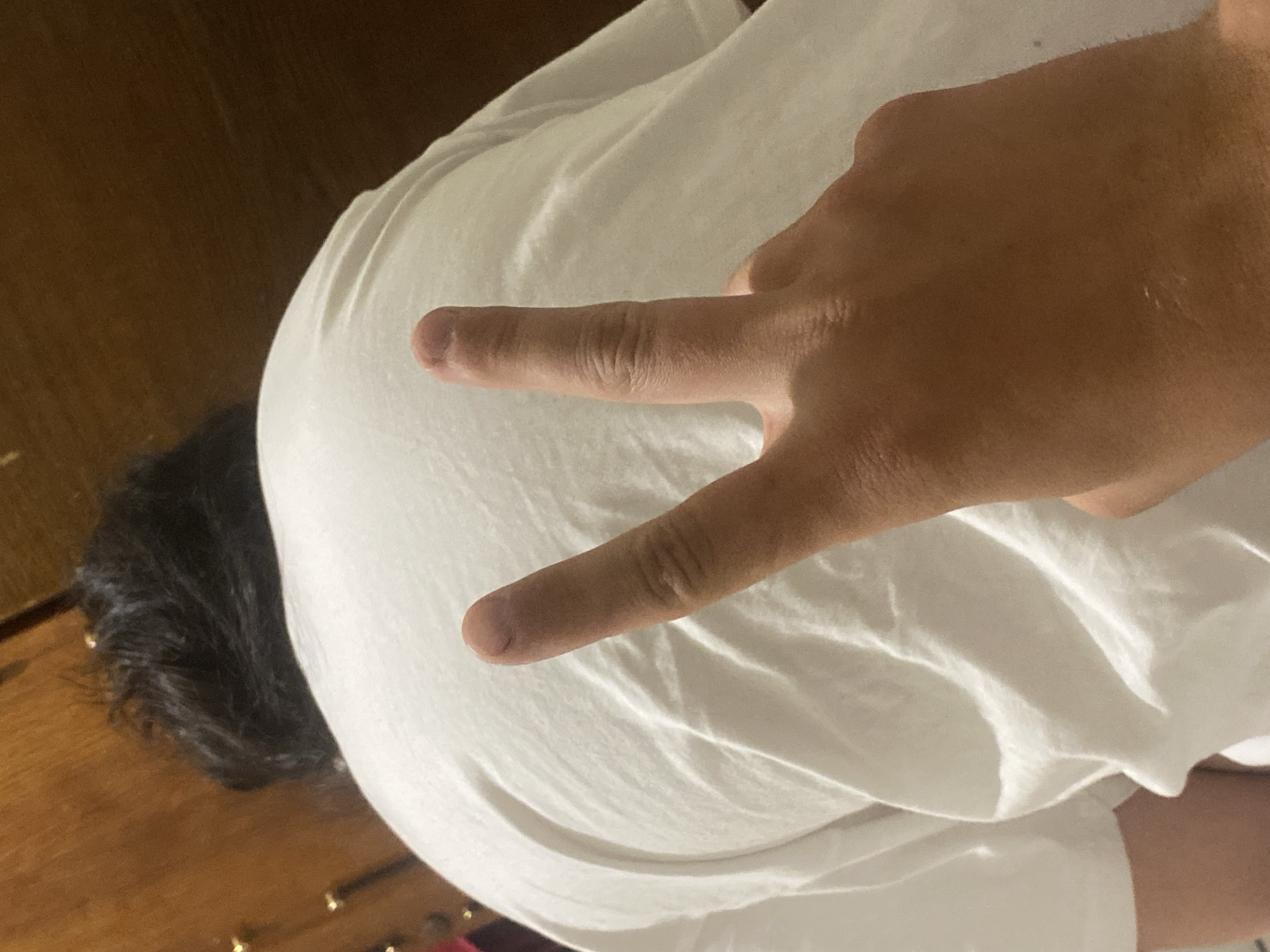 A person wearing a white t-shirt is lying on what appears to be a wooden floor. A hand is positioned above the person's back, displaying a peace sign. Part of the person's dark hair is visible near the edge of the frame. The location is not discernible from the image.