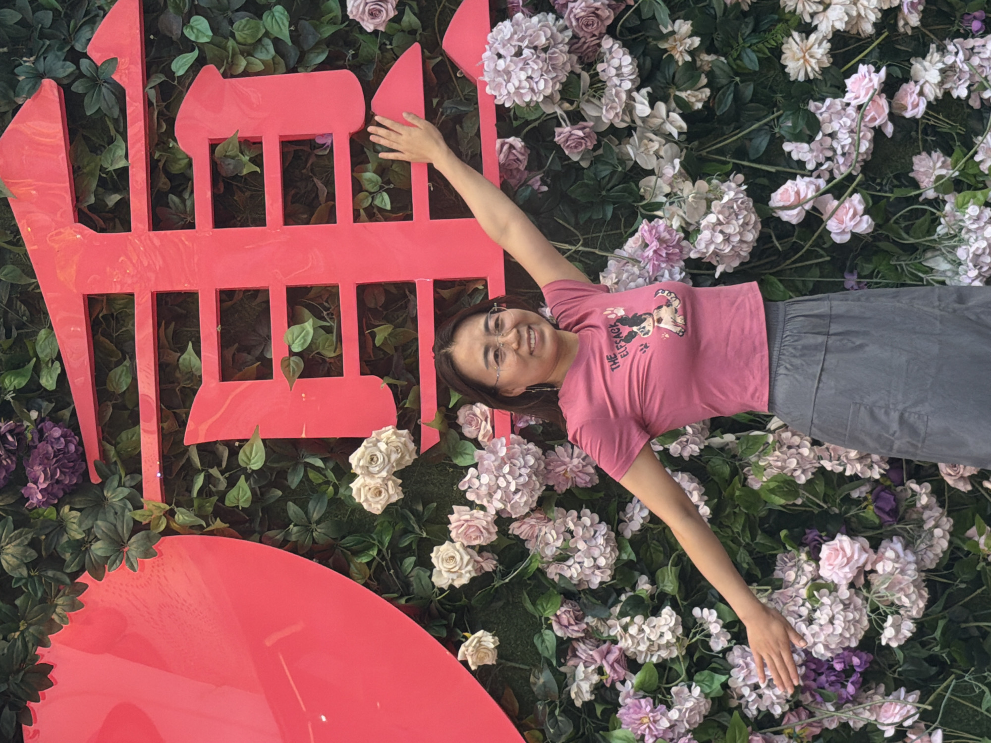 In Yuzhong District, China, a woman stands in front of a floral backdrop adorned with large, red Chinese characters. The woman, wearing a pink t-shirt with a graphic and a grey skirt, has her arms extended, one hand reaching towards the wall. The backdrop features an abundance of artificial flowers, including pink and white roses and hydrangeas, interspersed with green foliage. The red characters are bold and stylized, forming a significant visual element behind her. The lighting suggests an outdoor or well-lit indoor setting.