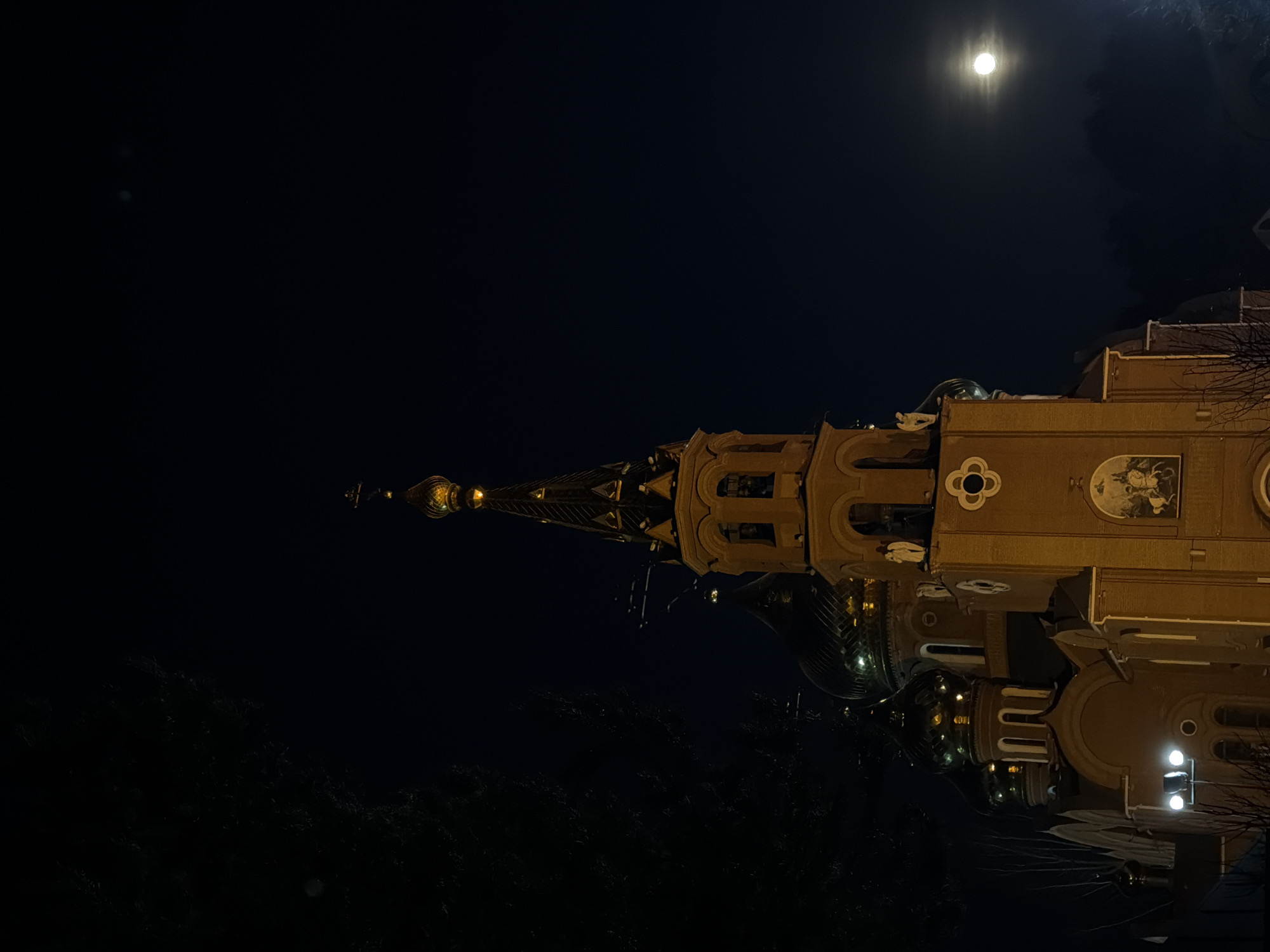 At night, a church is visible in Chaikovsky, Russia, illuminated by artificial lights. The church has a tall, pointed spire adorned with golden decorations and a dome. The church also has a building on the right side with arches, windows, and decorative elements. A full moon is visible in the upper-right corner. The environment is dark, with trees visible along the bottom, and clear night sky above.