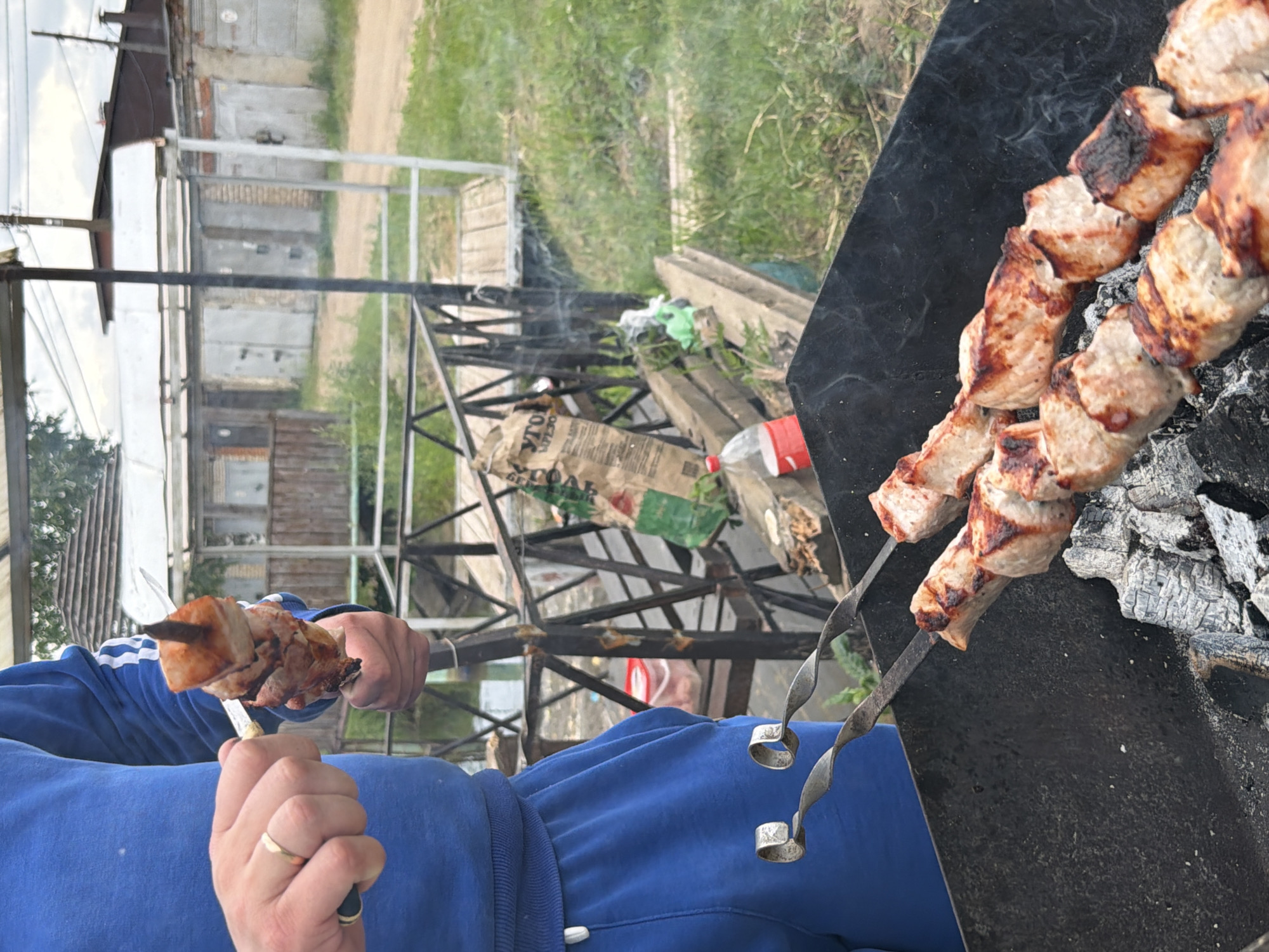 In Chaikovsky, Russia, a man wearing a blue tracksuit with white stripes on the sleeves is preparing shashlik, a popular Russian barbecue dish. He is holding a metal skewer with chunks of marinated meat, some of which are already being cooked over hot coals in a black metal grill. The grill is placed on a wooden surface, and smoke is rising from the coals and meat, indicating the cooking process is underway. In the background, there are several garages with gray metal doors, a wooden fence, and green grass, suggesting an outdoor residential area. A plastic bottle of red liquid and a paper bag are visible near the grill. The man is also wearing a gold wedding ring on his left hand.