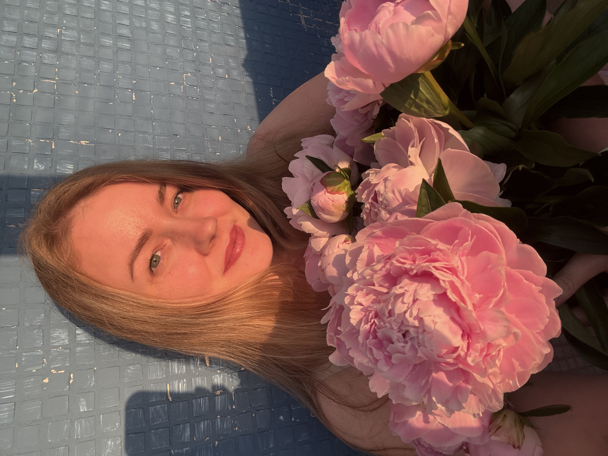 A young woman with blonde hair and blue eyes is holding a bouquet of pink peonies in front of a textured blue wall. The lighting suggests it is either sunrise or sunset, casting a warm glow on her face and the flowers. A shadow, possibly from her own profile or another person, is visible on the wall to her left. The background wall has a tiled or patterned surface. The woman is wearing a light-colored top. The image appears to be taken outdoors. No specific landmarks indicating the city of Chaikovsky, Russia, are visible in the image.