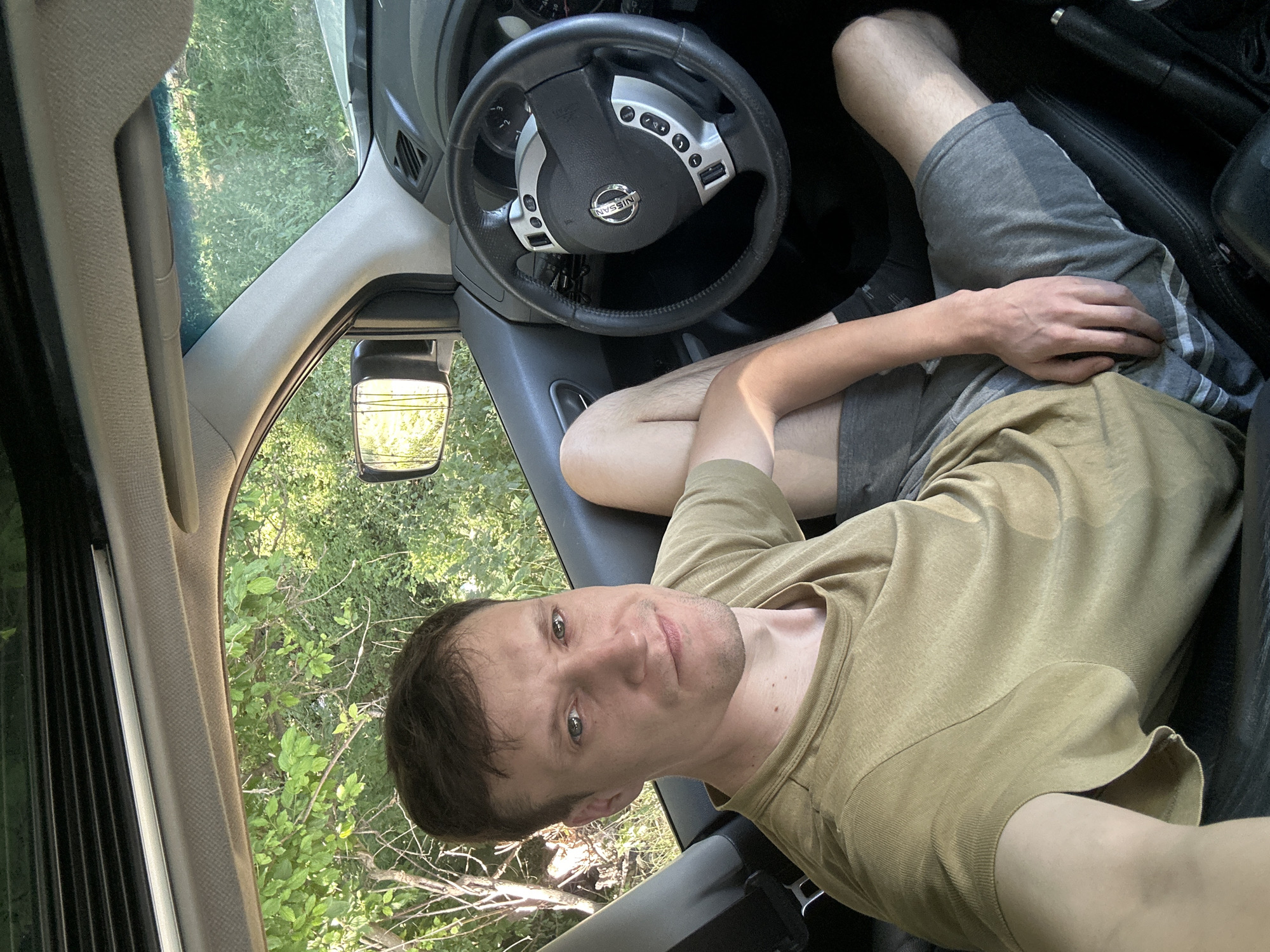 A man is sitting in the driver's seat of a car. He is wearing a tan t-shirt and grey shorts. The car's interior is visible, including the steering wheel and dashboard. The car is parked on a dirt road, surrounded by trees and foliage. The sun is shining, casting shadows on the scene. The man is looking at the camera with a neutral expression.