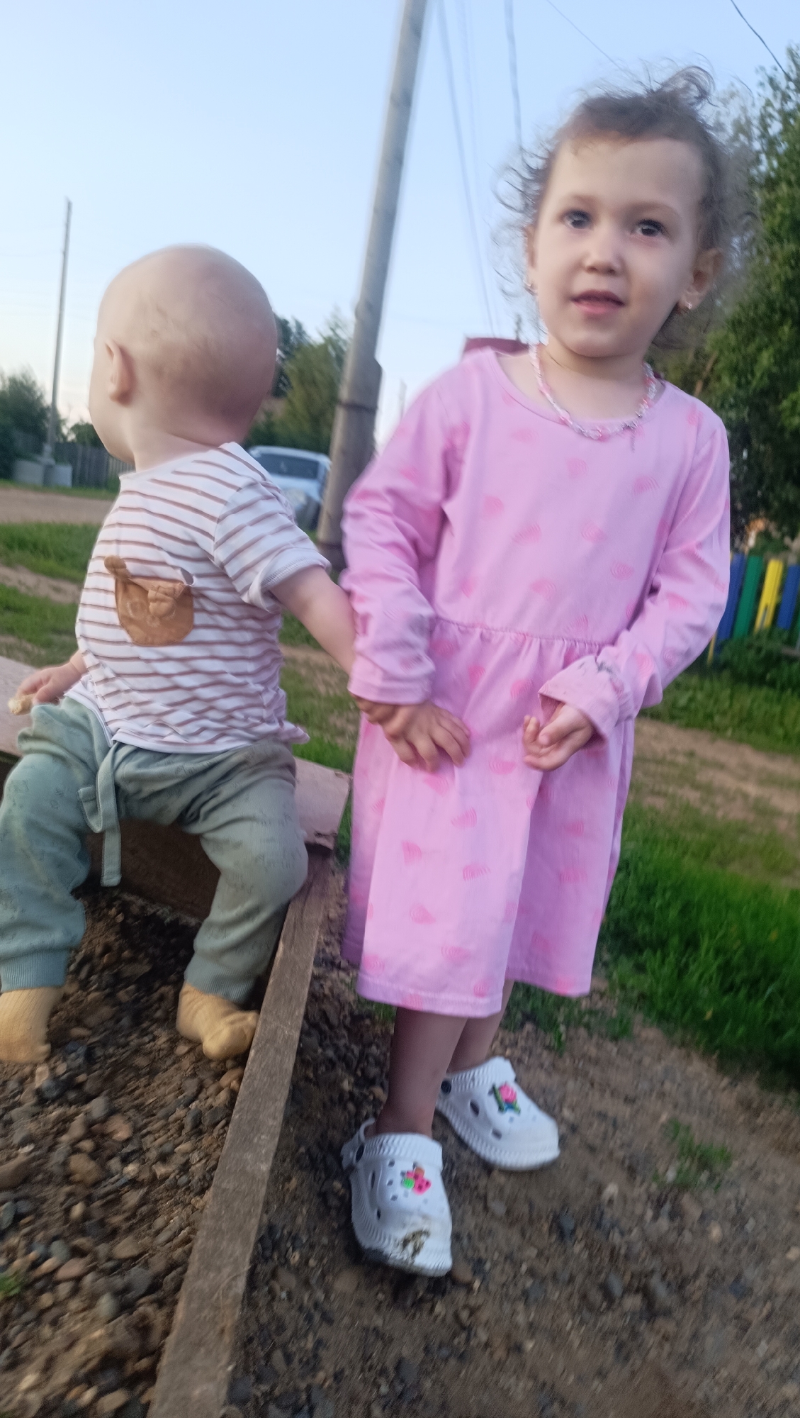 Two young children, a baby and a toddler girl, are standing next to each other outdoors, holding hands. The baby, facing left and seen from the side, is bald and wearing a striped t-shirt with a small brown bear embroidered on the chest and green pants. The toddler girl, facing the camera, is wearing a pink dress with a heart pattern and white Crocs with floral decorations.
They are standing on uneven ground with gravel and dirt. Behind them, a wooden structure is visible, and further back, a grassy area, a wooden fence with colorful elements, and trees can be seen. A utility pole and wires are also present in the background, suggesting a rural or semi-rural setting. A car is parked in the distance.
The lighting suggests it is late afternoon or early evening, with soft light and a pale sky. The overall mood appears calm and gentle. There is no visible text in the image. The location is identified as Karsha, Russia.