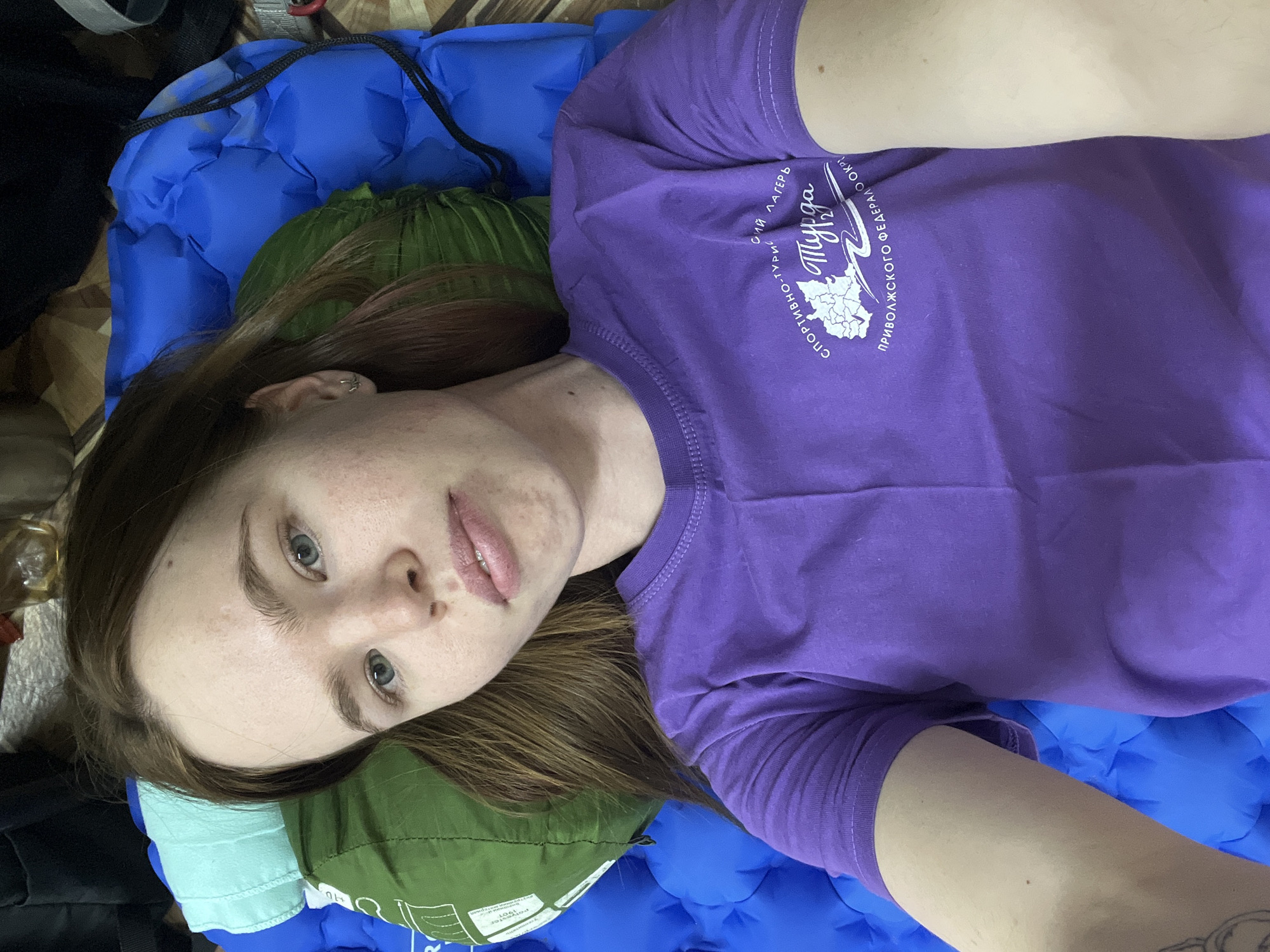 A woman is lying on an inflated blue camping mattress. She has light skin, light brown hair, and blue eyes. She is wearing a purple t-shirt with white text in Russian. The text includes the word "Myrda" and a map-like shape. The pillow is a green, tightly packed, cylindrical object. The woman's head is resting on the green pillow. Other dark-colored baggage is visible around the edges of the frame. There is no indication that the location is Berezniki, Russia.