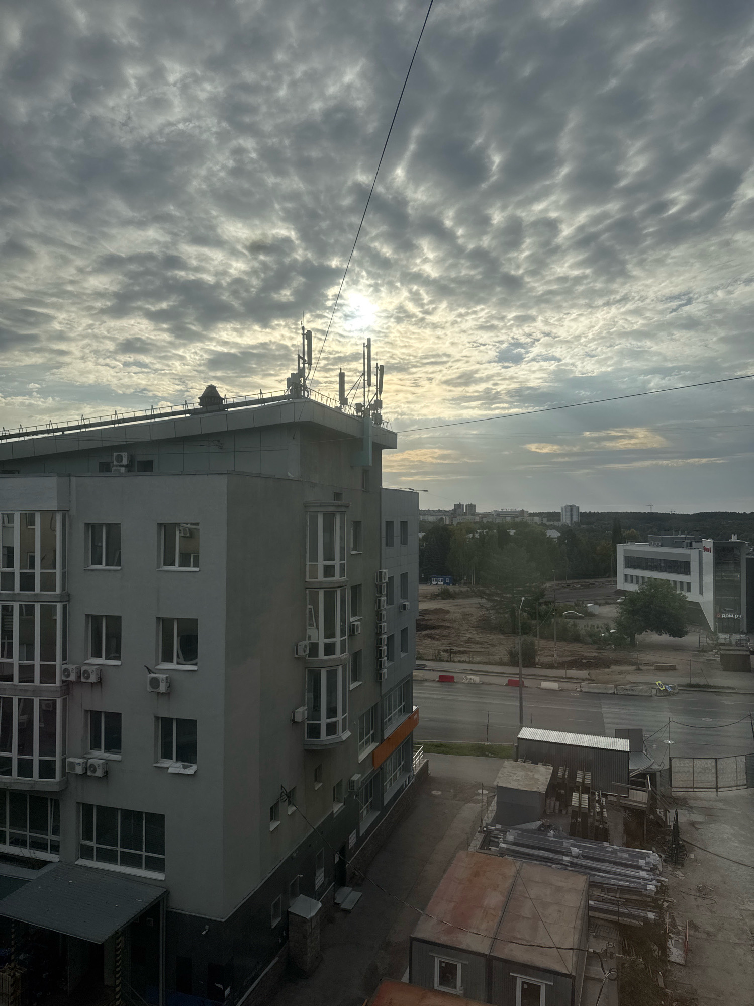 This is an exterior, eye-level, medium shot of a city street scene, taken during the daytime under a cloudy sky. The image is dominated by a modern, gray apartment building on the left, which features multiple windows and air conditioning units on its facade. The building's roof is equipped with multiple antenna arrays.
To the right of the apartment building, the view opens up to reveal a street with ongoing construction. Several temporary structures, possibly storage containers, are visible along with stacks of building materials, including pipes and metal beams. Beyond the construction area, a modern office building with a white facade and dark glass windows is visible. A sign on this building appears to read "дом.ру". Further in the distance, a treeline and other buildings can be seen, indicating a suburban or developing urban area. The sky is filled with dramatic, textured clouds with the sun peeking through in the upper center. A thin black wire stretches across the upper portion of the frame. The scene suggests a developing area within the city of Ufa, Russia. No people are visible in the frame.