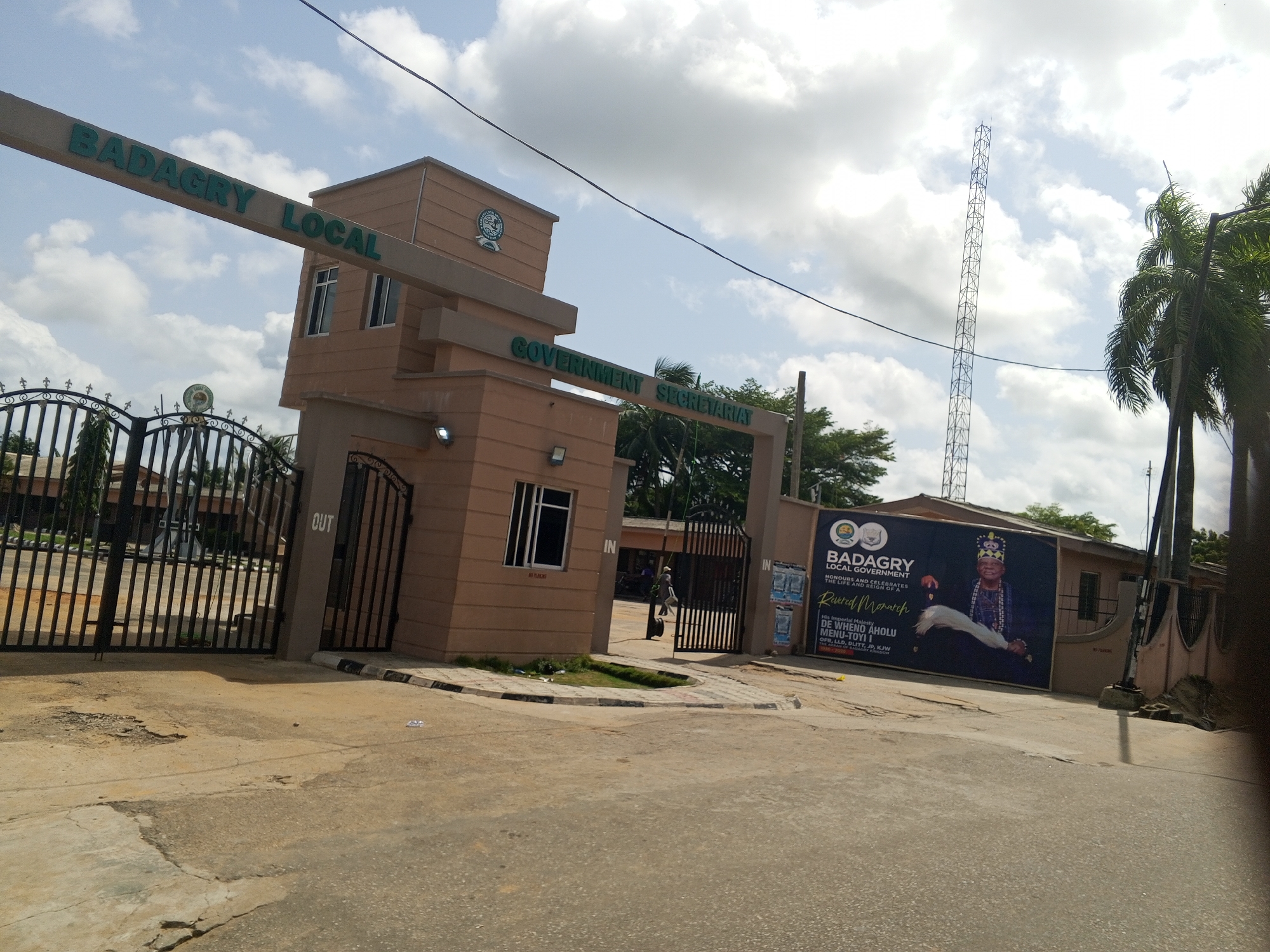 The image shows the entrance to the Badagry Local Government Secretariat in Badagry, Nigeria. The entrance features a gatehouse with signs reading "BADAGRY LOCAL GOVERNMENT SECRETARIAT". Metal gates are present for both entrance ("IN") and exit ("OUT"). A large banner is displayed near the entrance, depicting a man identified as His Imperial Majesty DE WHENO AHOLU MENU-TOYI I, OFR, LLD, DLITT, JP, KJW. A tall communication tower is visible in the background, along with palm trees and other vegetation. A person is seen walking through the entrance. The scene appears to be taken during daylight hours, with a cloudy sky above. The road leading to the entrance is paved.