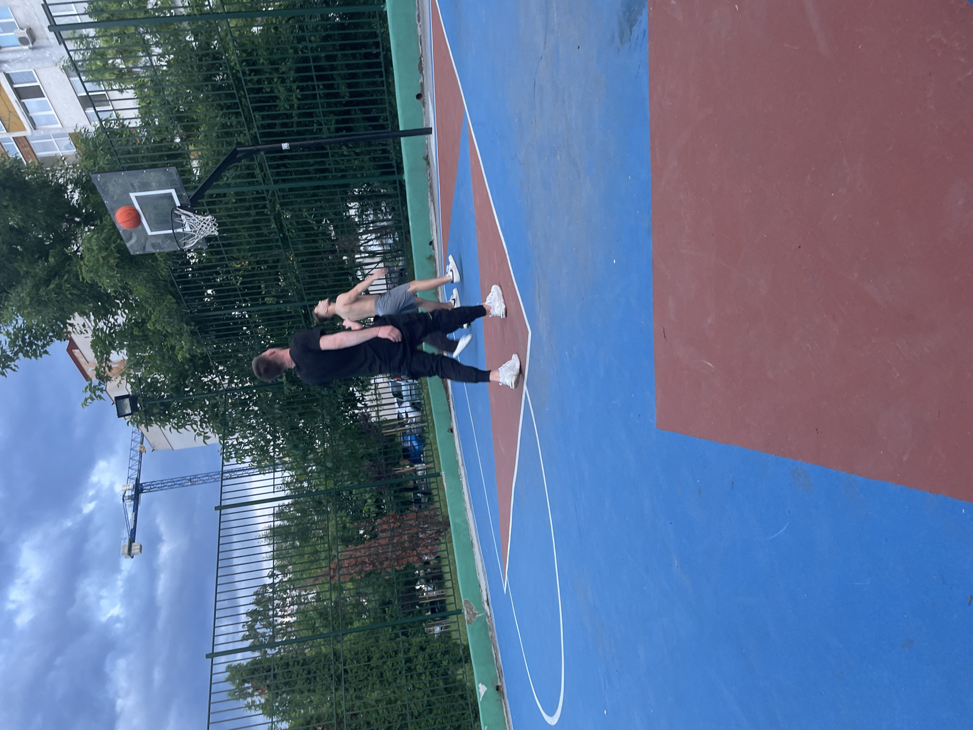 An outdoor basketball court, likely in Sofia, Bulgaria, is shown from a low-angle perspective. The court surface features distinct blue and reddish-brown sections with white boundary lines. The overall image appears to be rotated counter-clockwise by 90 degrees.
Two males are present on the court, engaged in a game of basketball. The person closer to the camera wears a black long-sleeved shirt, black pants, and white sneakers, standing with their back partially to the viewer. Further down the court, another male, shirtless and wearing grey shorts and white sneakers, faces the first person. An orange basketball is clearly visible within the net of the basketball hoop, indicating a successful shot or the ball passing through. The hoop itself has a clear backboard.
The court is surrounded by a green metal fence and lush green trees. In the background, an apartment building with visible windows and an air conditioning unit stands tall, and a large blue construction crane is silhouetted against a cloudy, overcast sky. The lighting suggests it is daytime, possibly late afternoon, given the soft, diffused illumination. The court surface shows signs of wear, with some scuff marks and discoloration. No discernible text is visible on any objects or surfaces.