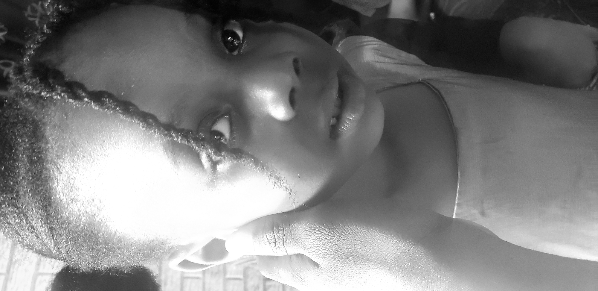 This is a close-up, high-contrast black and white photograph depicting a young female child, understood to be situated in Narayi, Nigeria. The child, with dark, textured hair styled into prominent side buns or puffs, has several strands falling across her forehead and right cheek. Her eyes are wide open, dark, and look directly upwards and slightly towards the viewer, reflecting a bright light source. Her nose is prominent, and her mouth is slightly open, revealing some lower teeth and upper incisors, suggesting a natural or curious expression. A hand, with distinct skin texture visible, is pressed against the child's left cheek and ear, with the thumb and fingers clearly delineated. She is wearing a light-colored garment, possibly a t-shirt or tank top, visible around her neck and chest area. The background on the upper left consists of a light-colored brick wall with visible mortar lines, while the rest of the background is indistinct and darker.