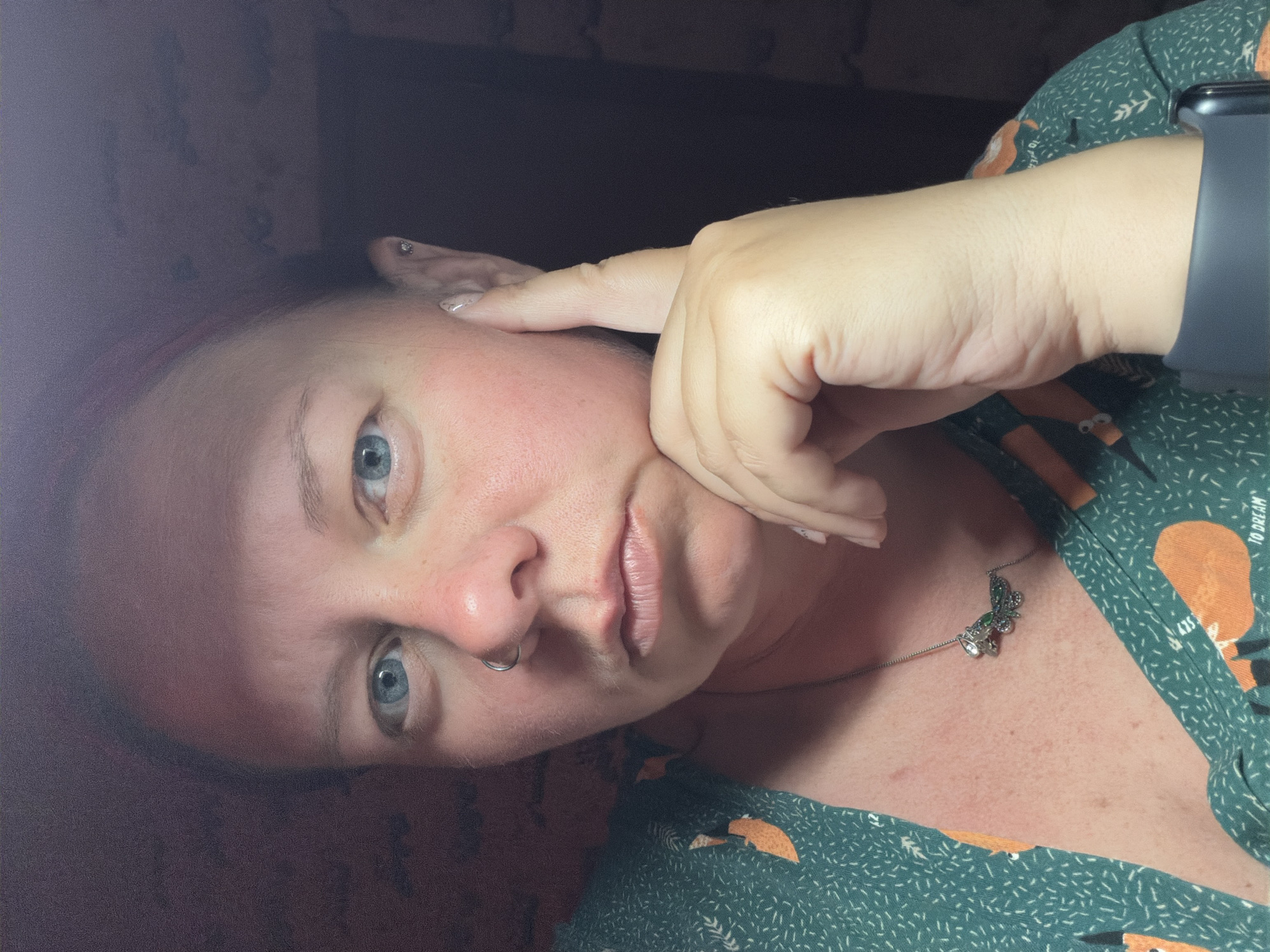 A pale-skinned person with blue eyes and a nose ring is visible, turned at an angle to the camera. They are wearing a green patterned shirt with a necklace featuring a butterfly-shaped pendant. The person's right hand is raised to their face, with one finger touching the temple. The background is dark and indistinct. The image location is unknown.