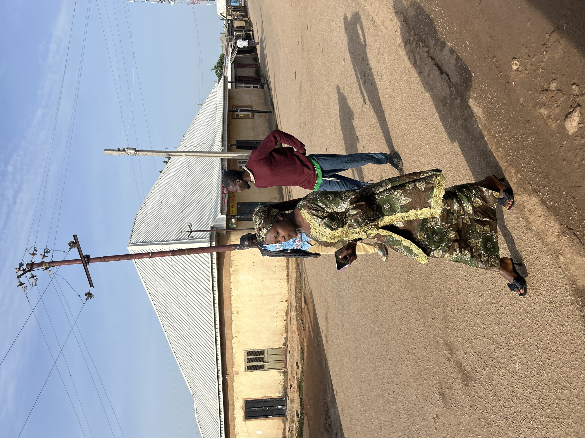 In Jos, Nigeria, a woman in a green and gold patterned dress and headscarf walks along a dusty, unpaved street. She holds a dark-colored smartphone in her right hand. Behind her, two men walk in the same direction; one in a blue shirt and light-colored trousers, the other in a maroon long-sleeved shirt and blue jeans. The buildings lining the street are simple, single-story structures with corrugated metal roofs, painted in muted yellow and beige tones, featuring closed wooden doors and windows. Electrical poles with multiple wires are visible overhead. The scene is illuminated by natural daylight, casting long shadows on the ground.